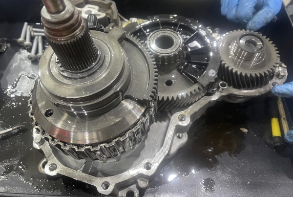 Close-up of a partially disassembled metal gearbox with gears and bearings on a workbench, with gloved hands holding one side component.