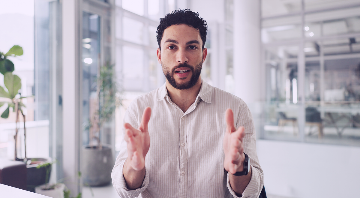 Man with curly hair and beard speaking and gesturing with hands in a modern office setting.