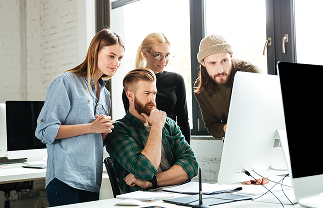 Four young professionals gathered around a computer screen in a bright office, engaged in collaborative work.