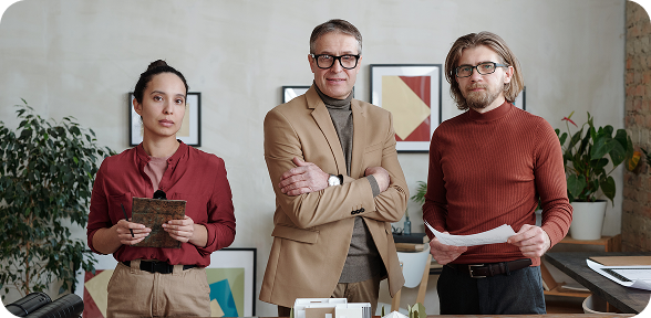 Three professionals standing in an office; two men and one woman holding papers and a clipboard.