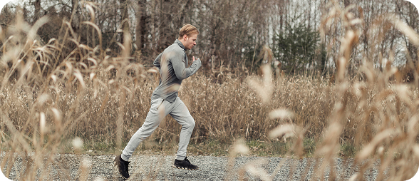 Young man jogging on a gravel path surrounded by tall dry grass and leafless trees in a natural outdoor setting.
