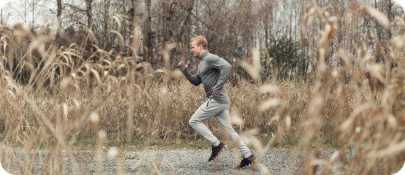 Man running on a gravel path surrounded by tall dry grass and leafless trees in autumn.