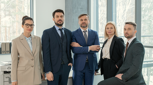 Five diverse business professionals in suits standing and sitting in a modern office with large windows.