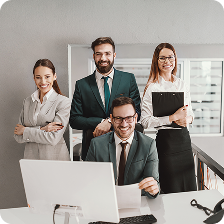Four business professionals smiling in an office, with one seated at a desk using a computer and the others standing behind.