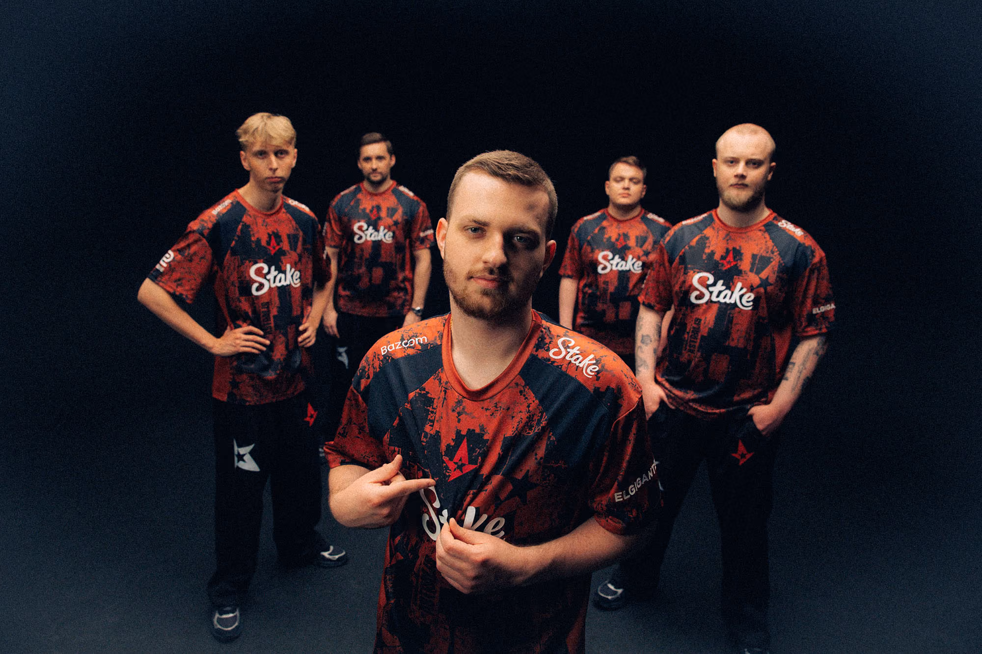Wide group portrait of five Astralis players in red-and-black Simon Wick jerseys against a dark backdrop, with one player showcasing the crest.