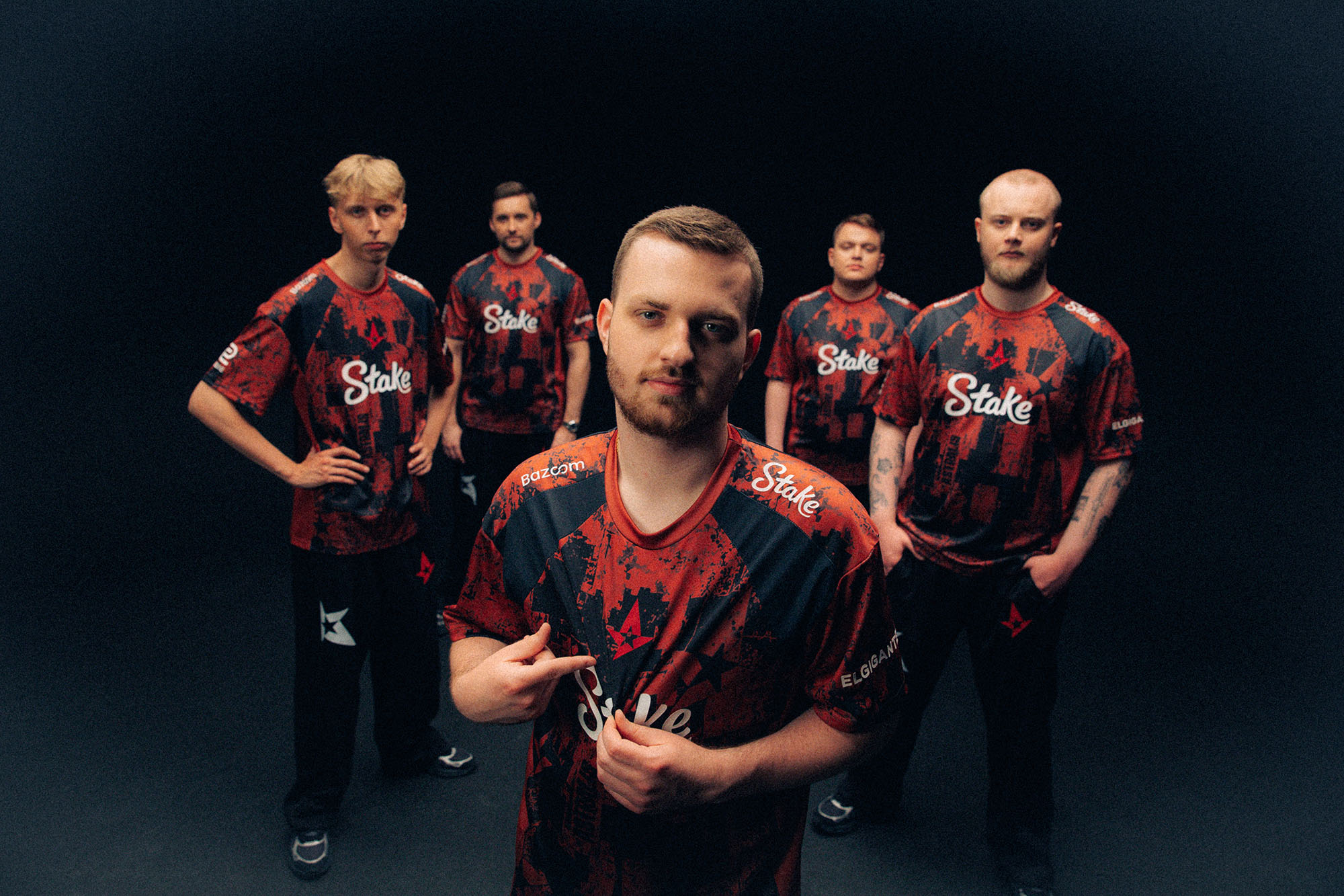 Wide group portrait of five Astralis players in red-and-black Simon Wick jerseys against a dark backdrop, with one player showcasing the crest.