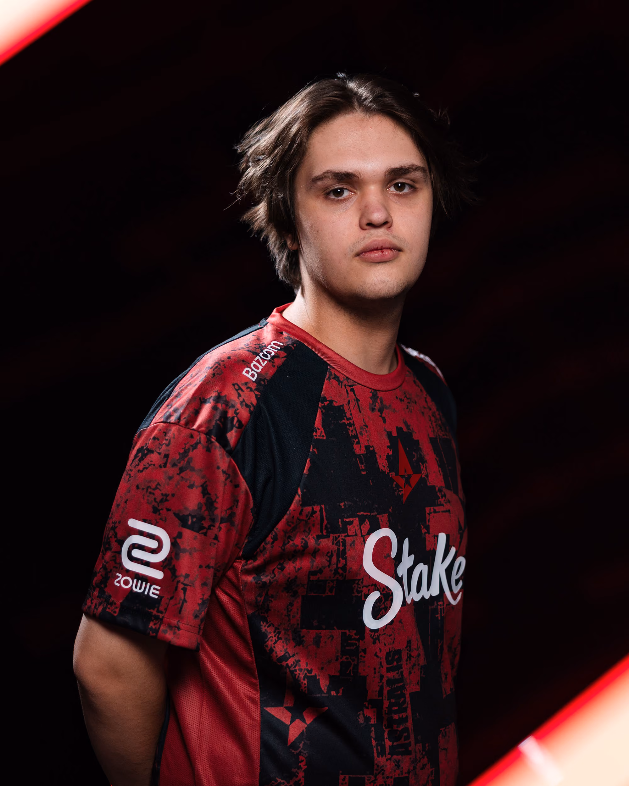 Young man with blonde hair wearing a black and red 'Stake' shirt against a dark background.