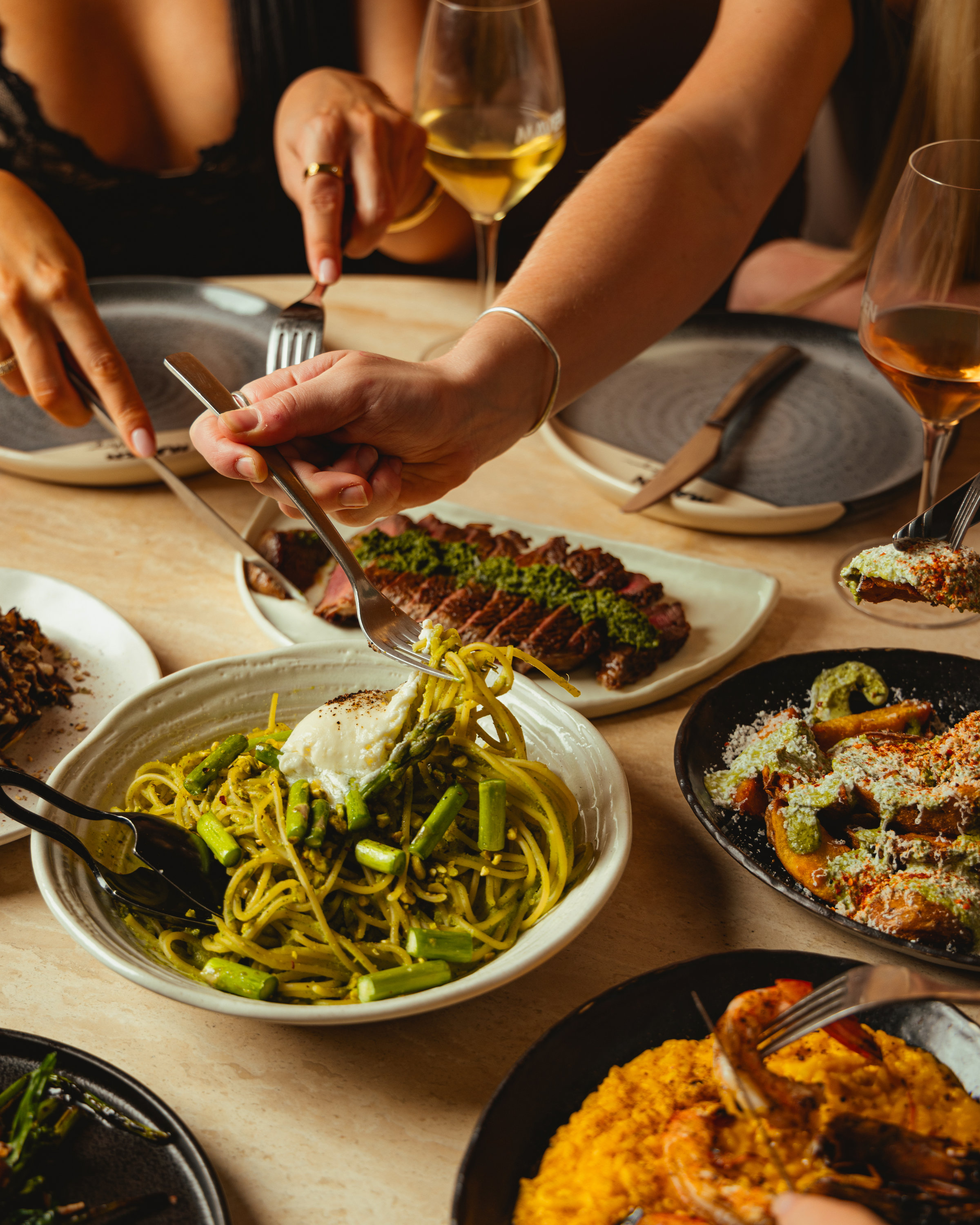 Guests sharing pesto spaghetti with burrata and asparagus, sliced steak with salsa verde, and wine at Maven