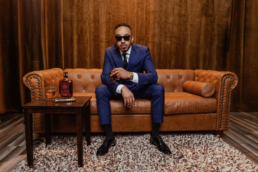 Man in navy suit and sunglasses sitting on leather sofa with bourbon