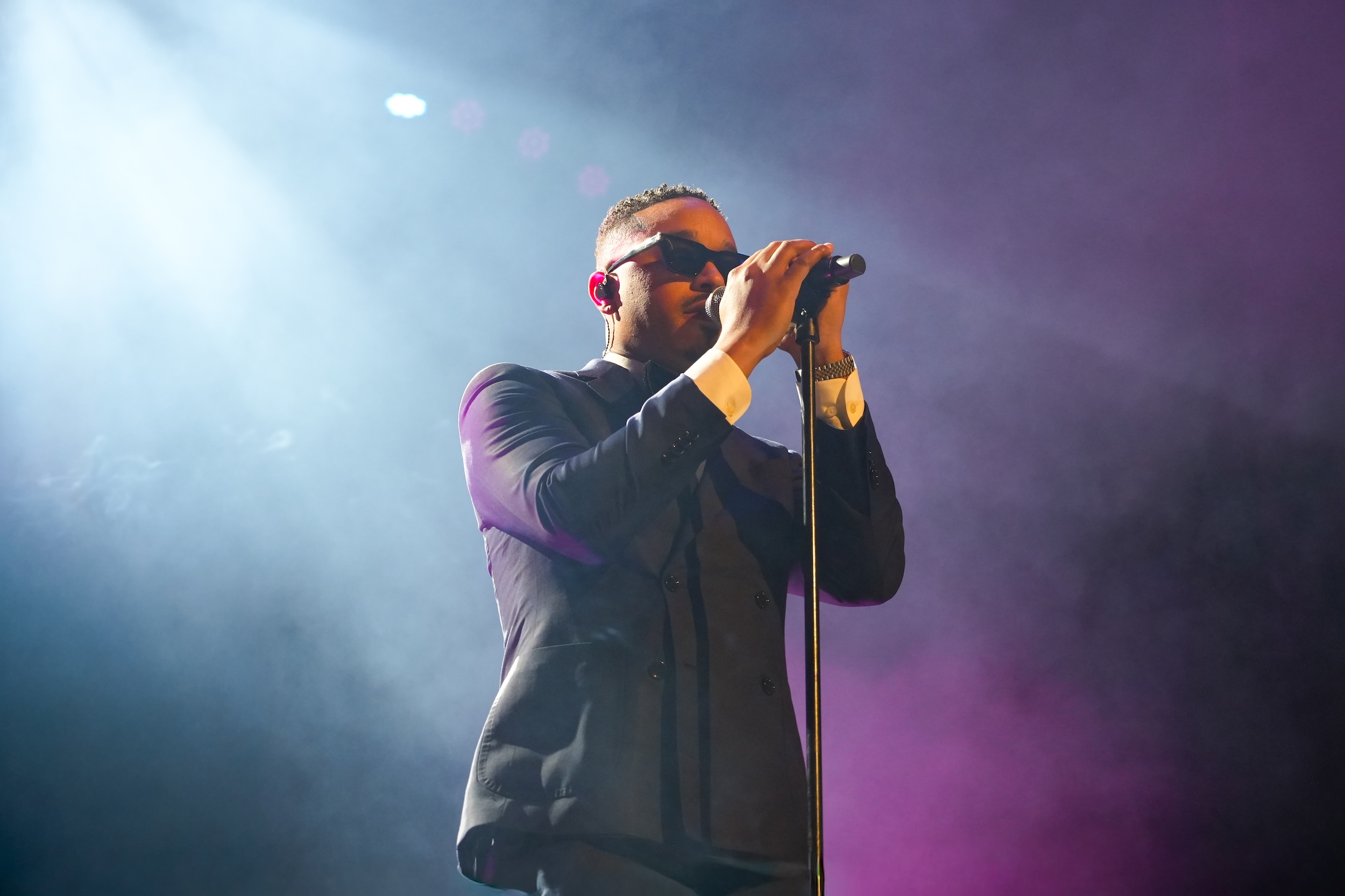 Male performer singing passionately on stage with sunglasses and microphone in colorful lighting.
