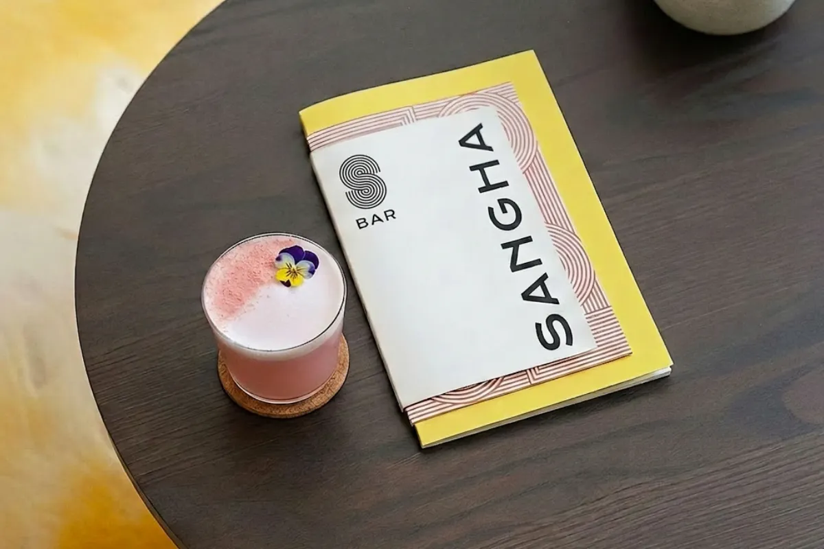 A pink cocktail garnished with an edible flower on a coaster next to a yellow and white menu titled 'SANGHA' on a dark wooden table.