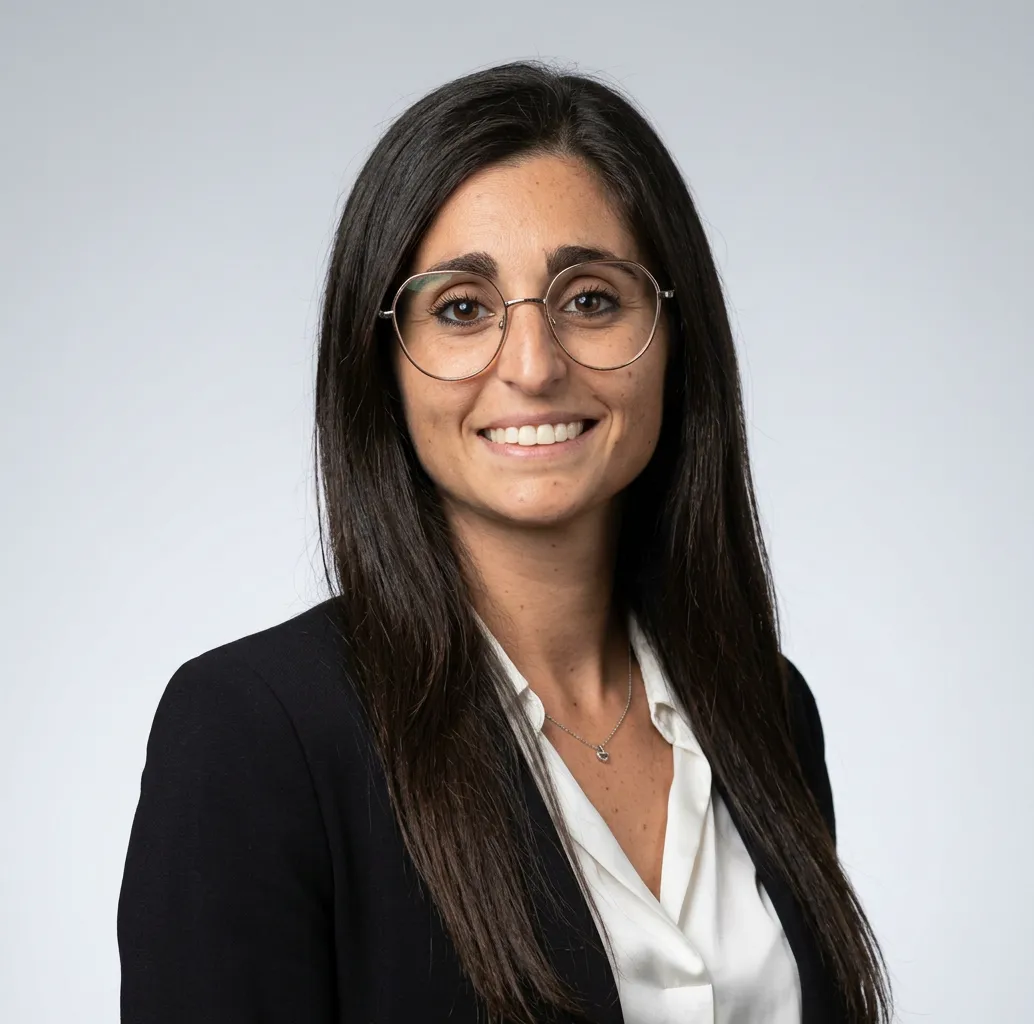 Smiling woman with long dark hair wearing round glasses, a white blouse, and a black blazer against a light gray background.