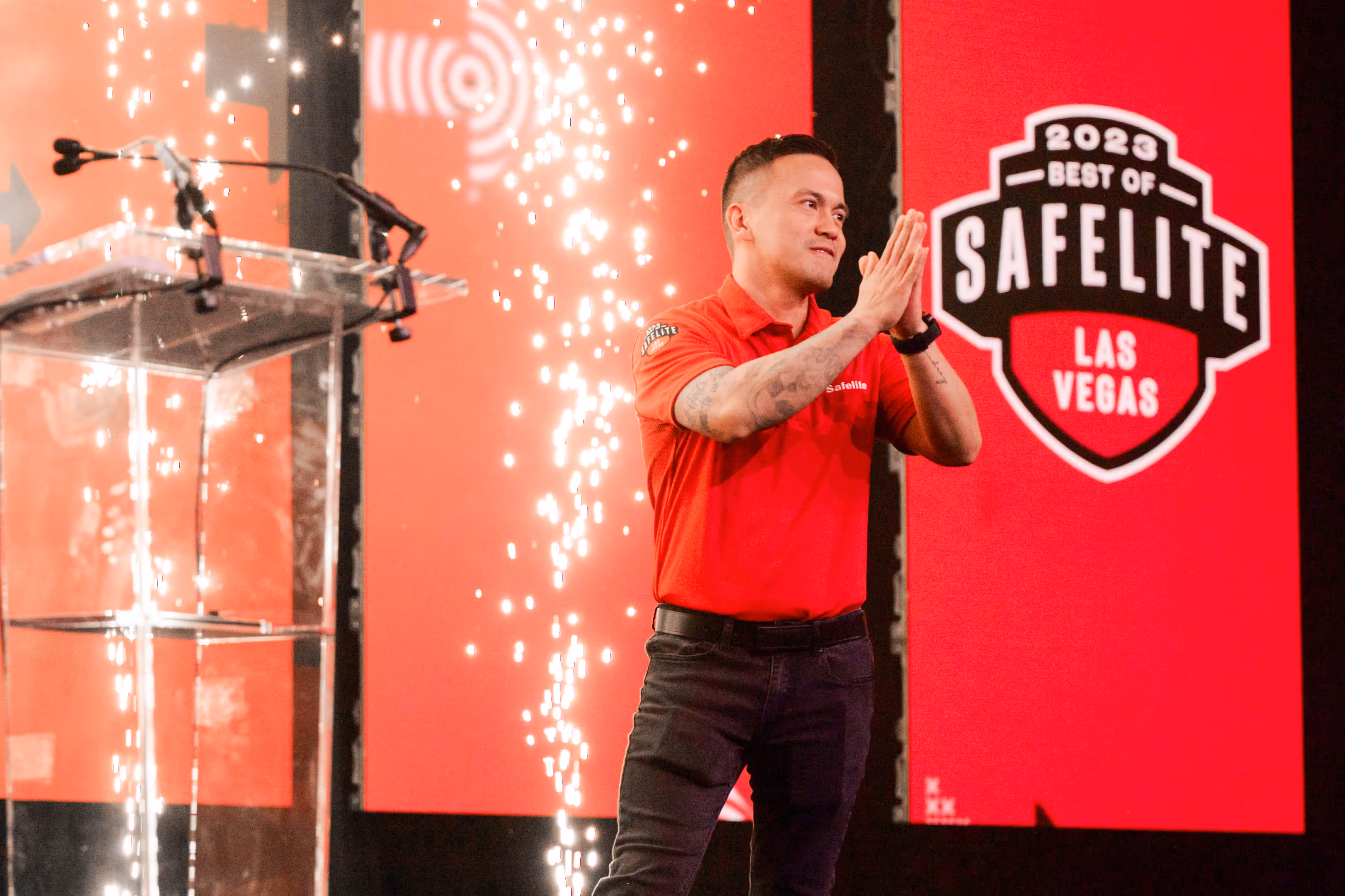 Man in a red Safelite shirt clapping on stage with pyrotechnics and a '2023 Best of Safelite Las Vegas' sign in the background.