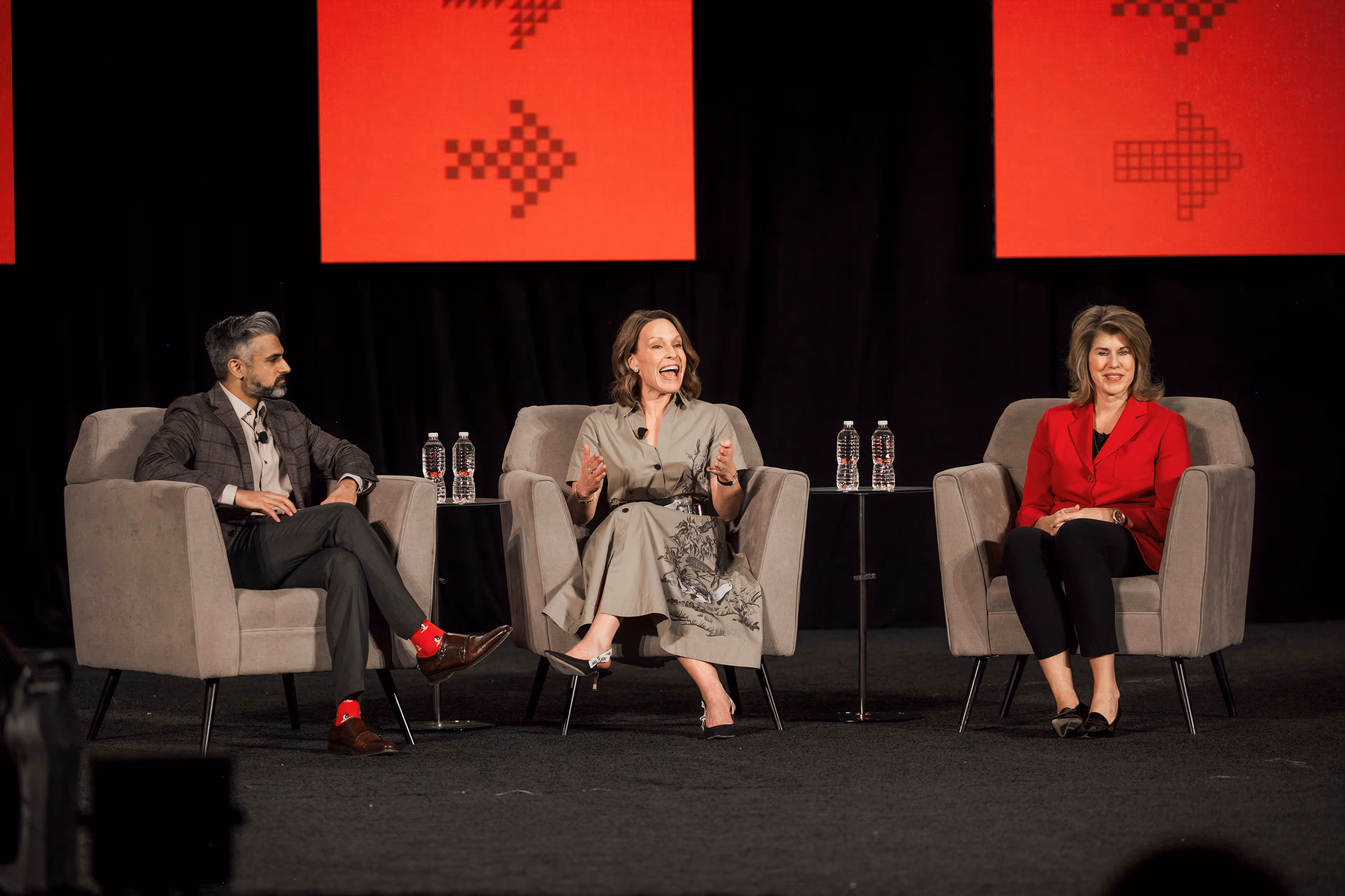 Three people sitting on stage in armchairs engaged in a panel discussion with two small tables holding water bottles between them.