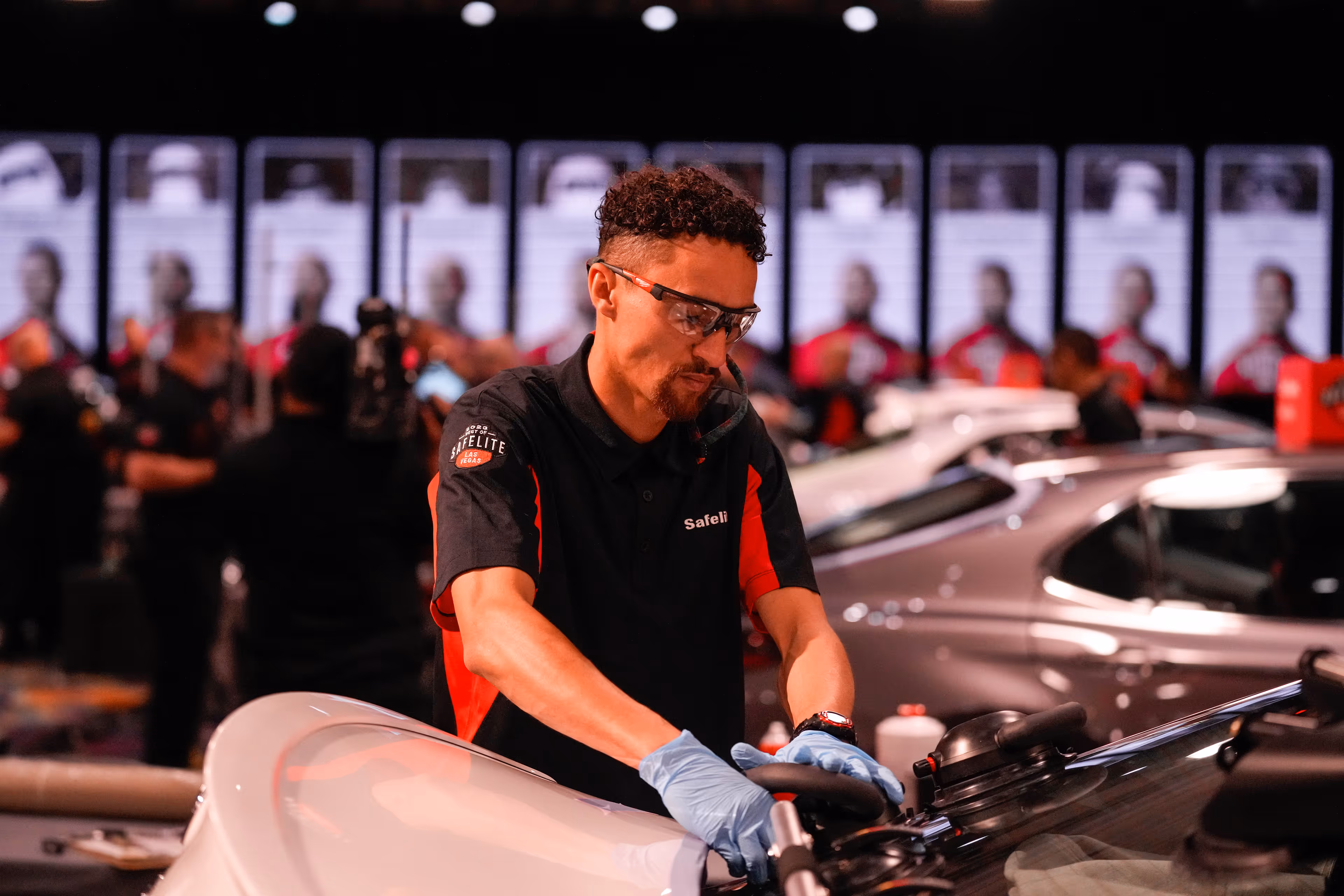 Man wearing safety glasses and gloves working on a car hood in a workshop with other cars and people blurred in the background.
