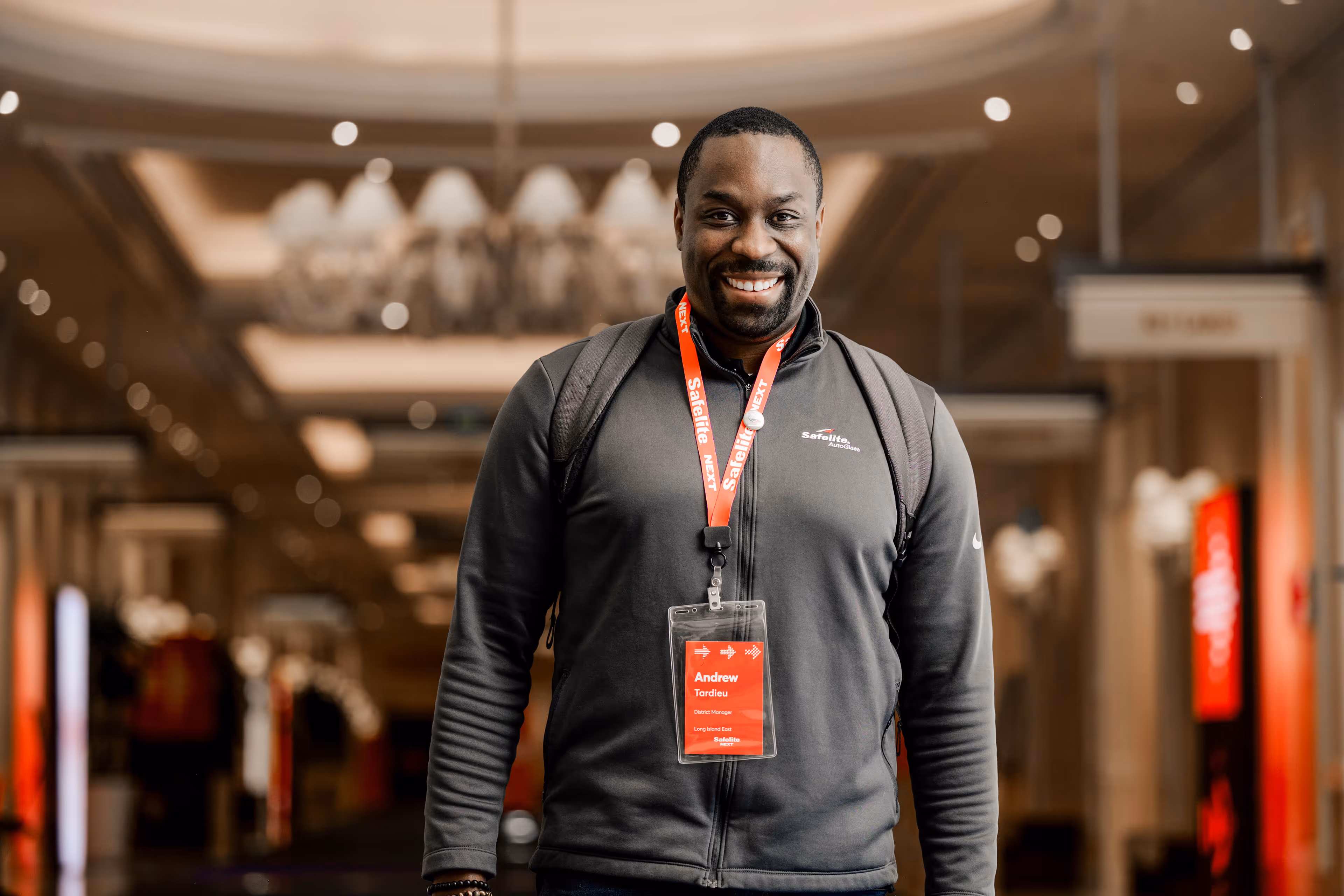 Smiling man wearing a gray Safelite jacket and a red event badge with the name Andrew Tardieu, standing indoors with blurred background.