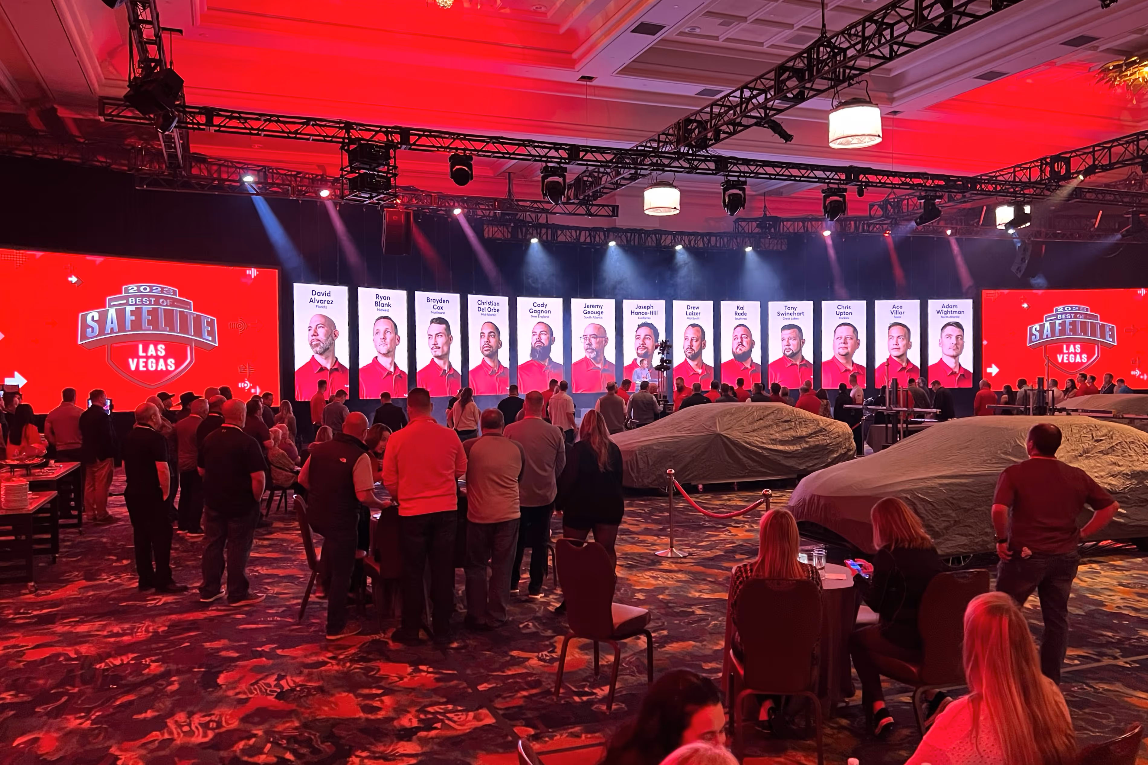 Audience watching a stage with multiple vertical screens showing portraits and names of men in red shirts for the 2023 Best of Safelite Las Vegas event, with covered vehicles in front of the stage.