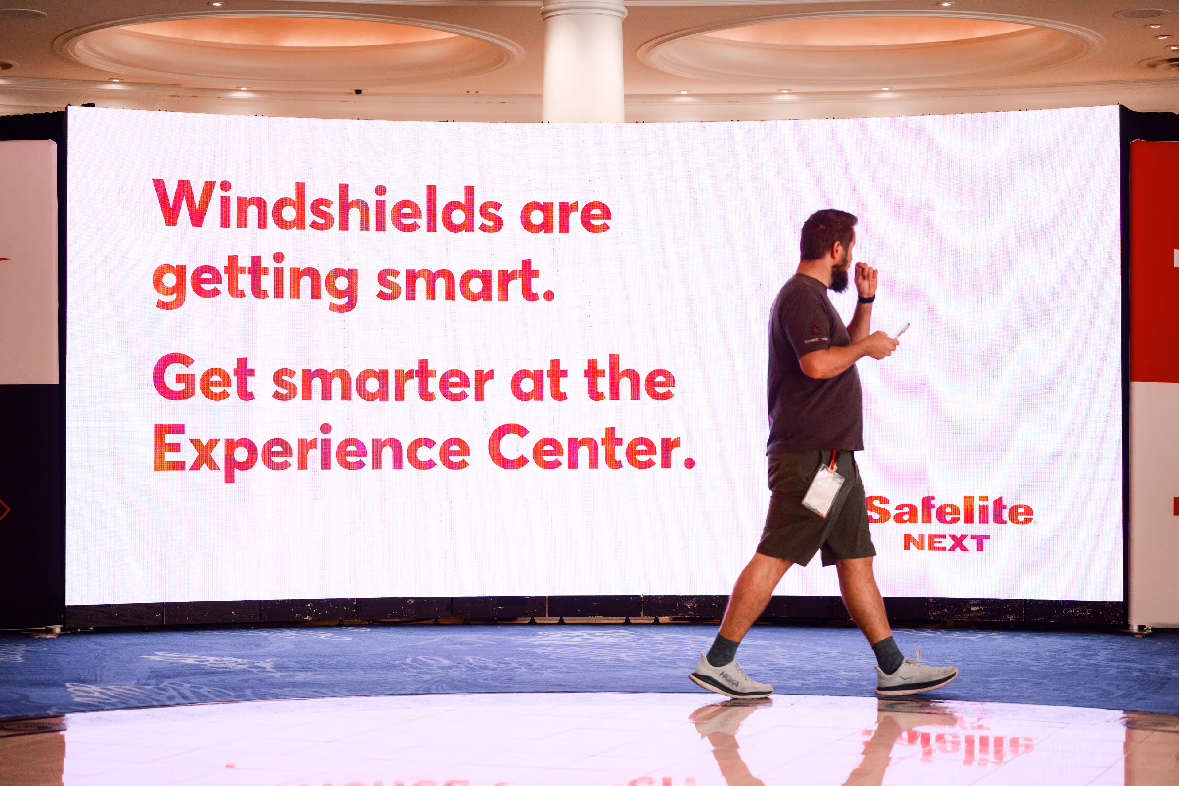 Man walking in front of a large digital display that reads: Windshields are getting smart. Get smarter at the Experience Center. Safelite NEXT.
