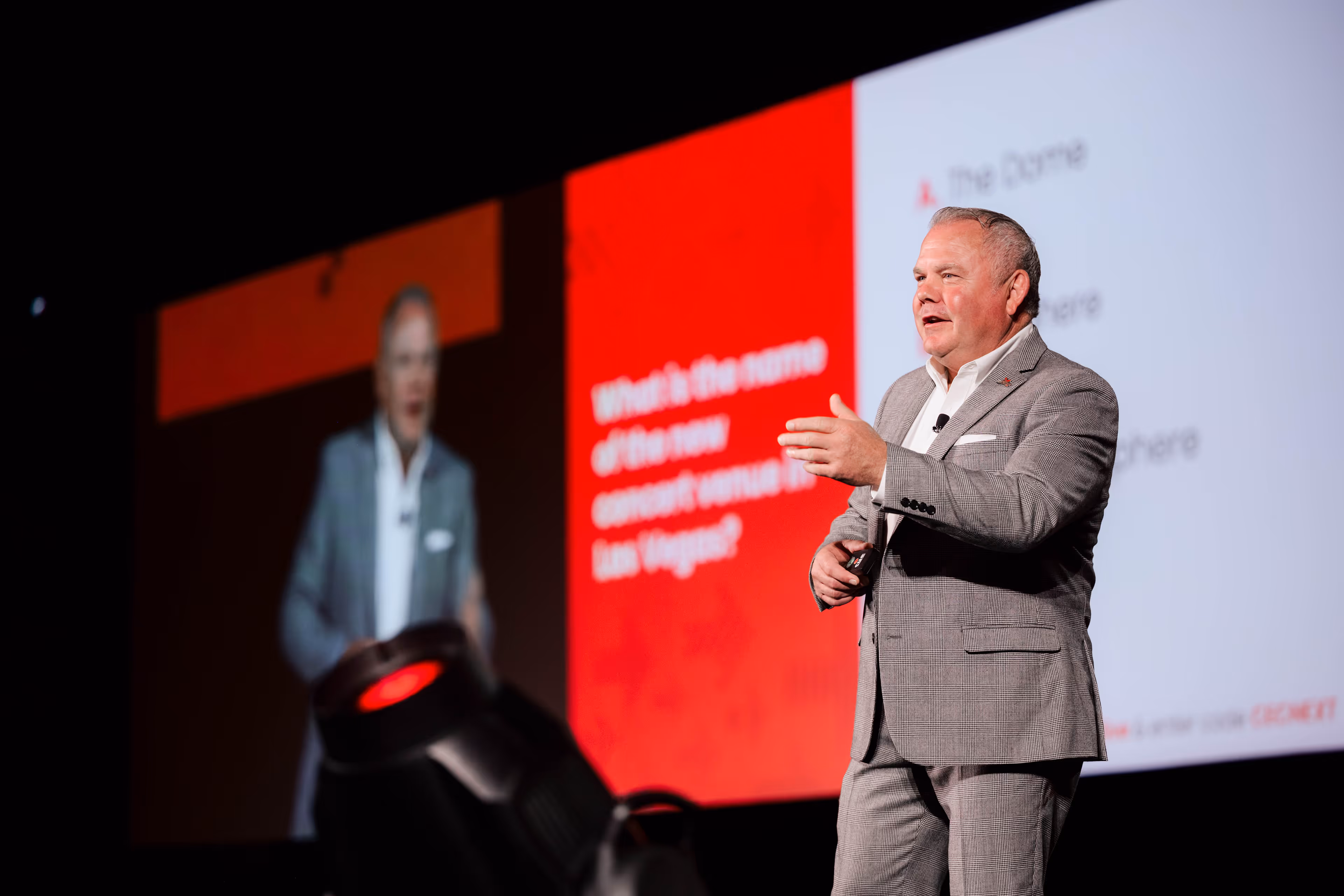 A man in a gray suit giving a presentation on stage with a large red and white screen behind him displaying text about a new concert venue in Las Vegas.