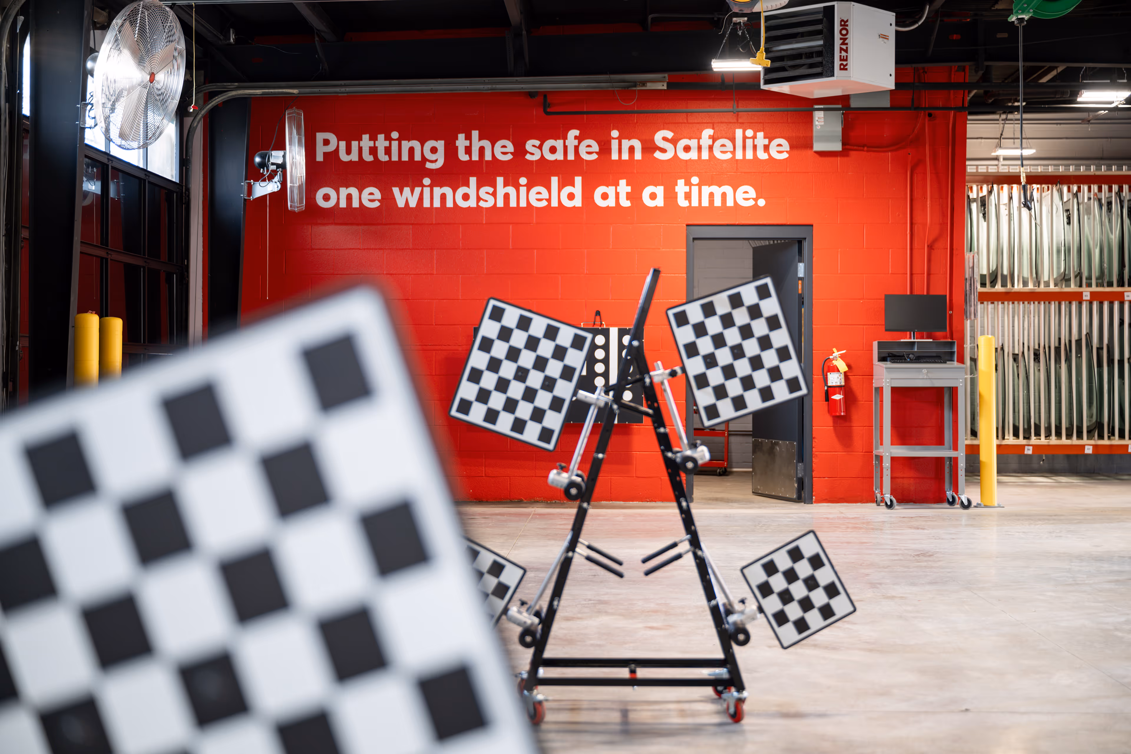 Red wall inside an auto repair facility with the white text 'Putting the safe in Safelite one windshield at a time' and a checkerboard calibration tool in the foreground.