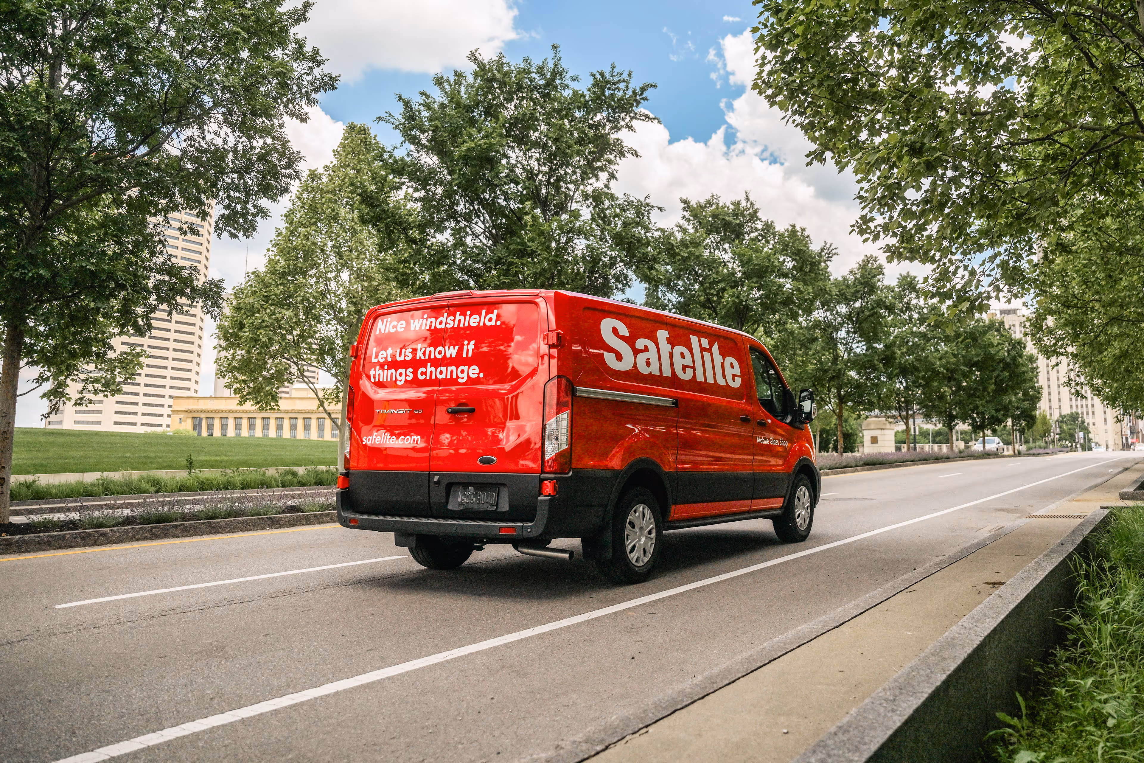 Red Safelite service van driving on a city street lined with trees and buildings in the background.