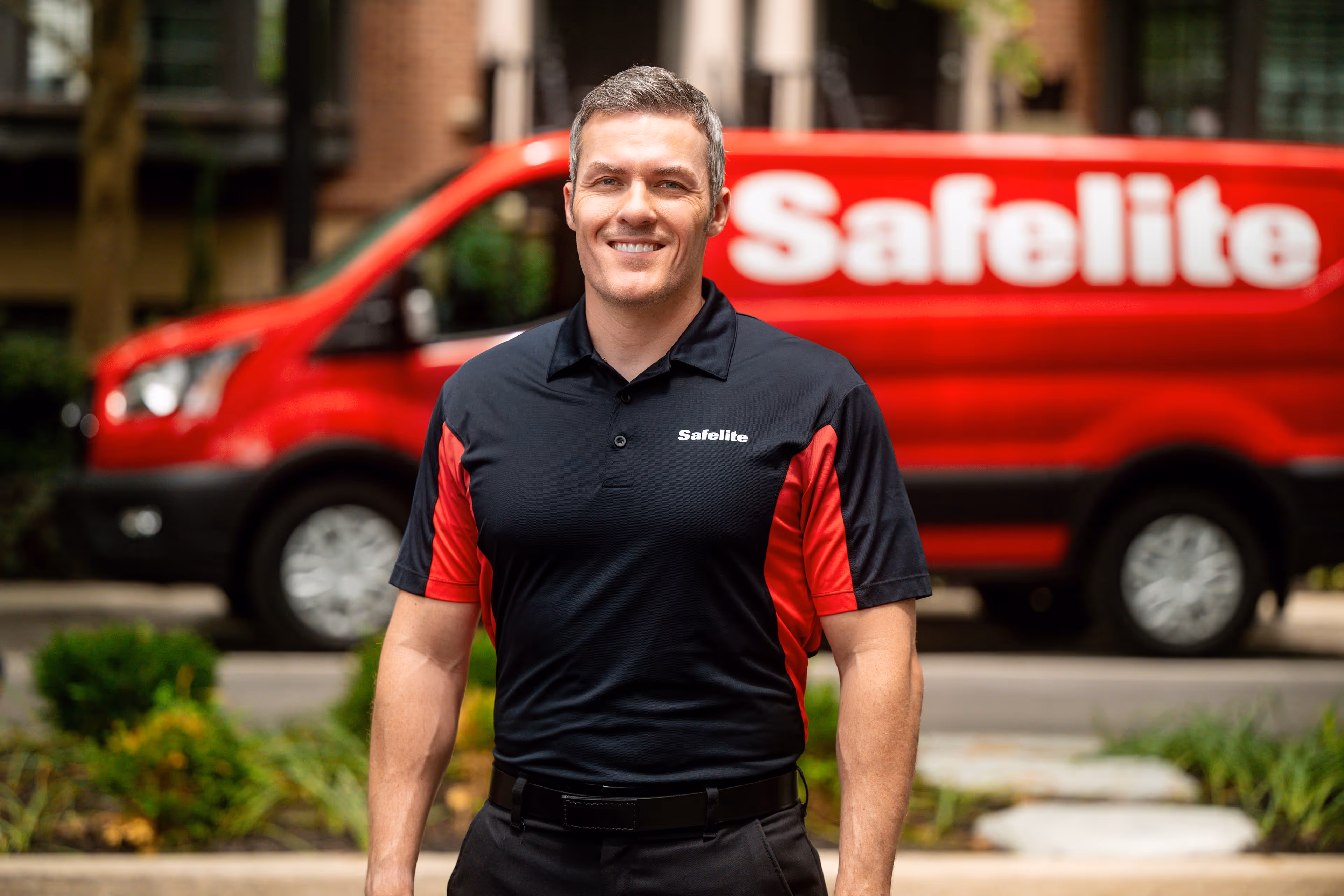 Smiling man wearing a black and red Safelite uniform shirt standing in front of a red Safelite service van.