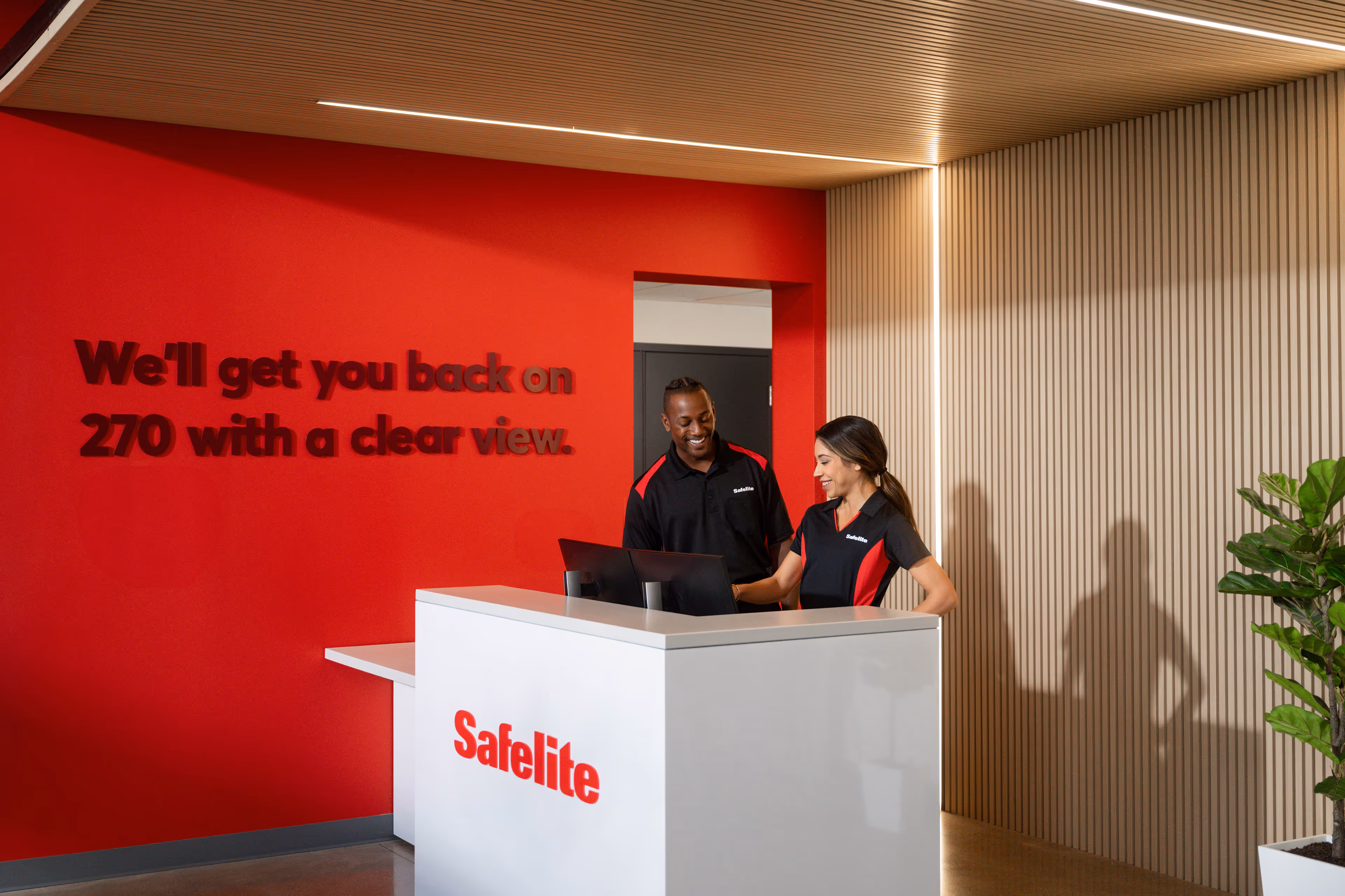Two Safelite employees in black and red uniforms smiling and working behind a white reception desk in a modern office with red and beige walls.