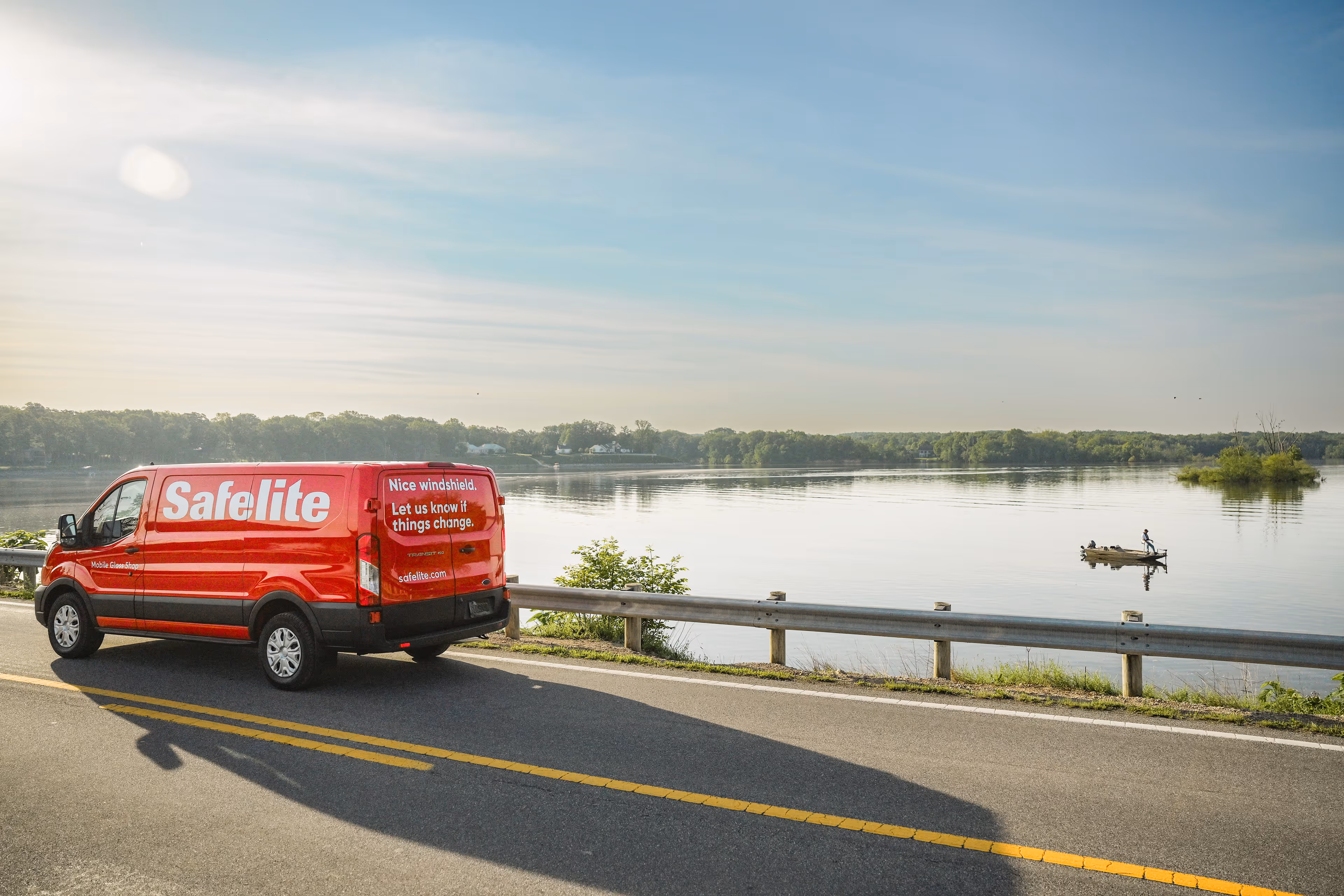 Red Safelite van parked on a road beside a calm lake with a small boat in the water and a tree line in the distance under a clear sky.