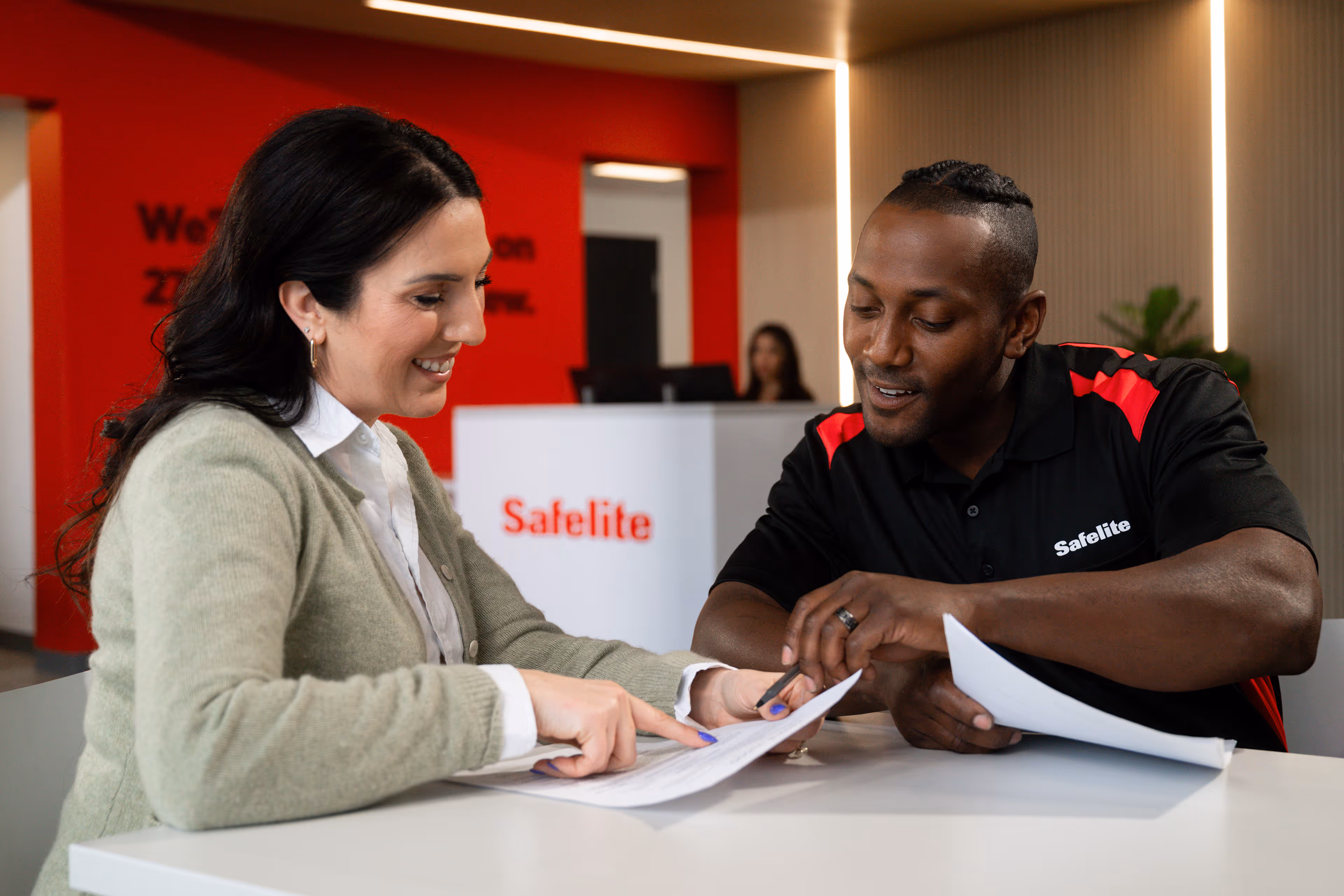 Smiling woman and Safelite employee review documents together at a white table in a Safelite office lobby.