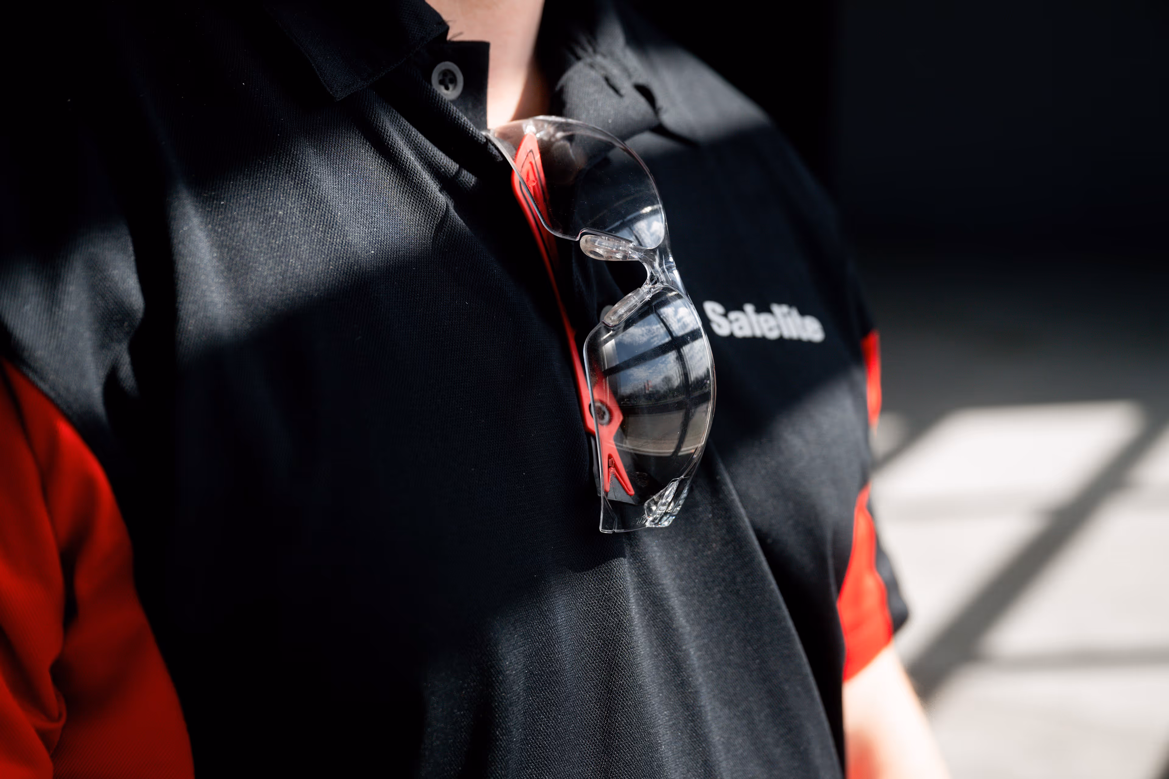 Close-up of a person wearing a black and red polo shirt with the word 'SafeFlo' and clear safety glasses hanging from the collar.