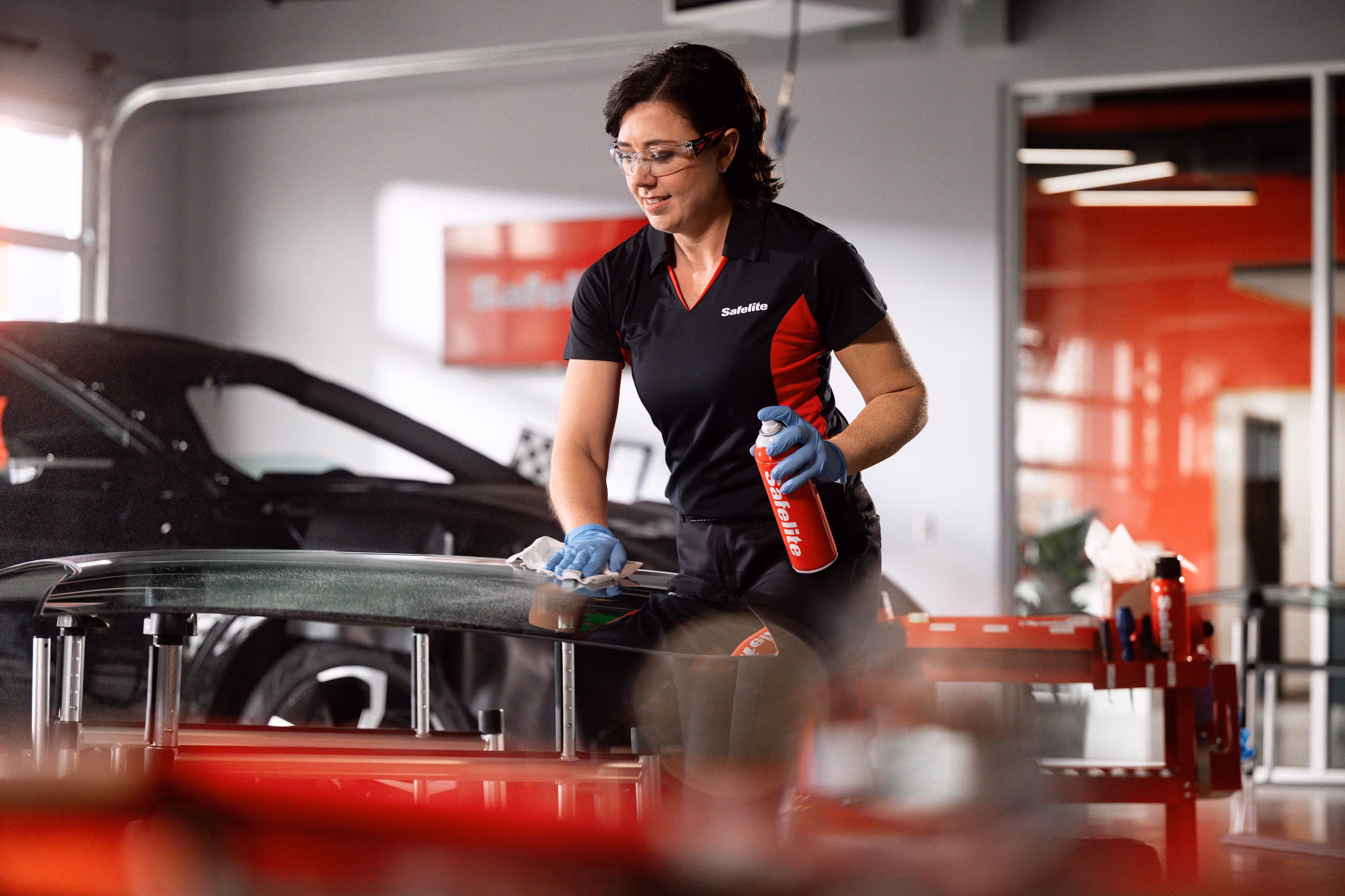 Woman technician wearing Safelite uniform and safety glasses cleaning a car windshield in a garage.