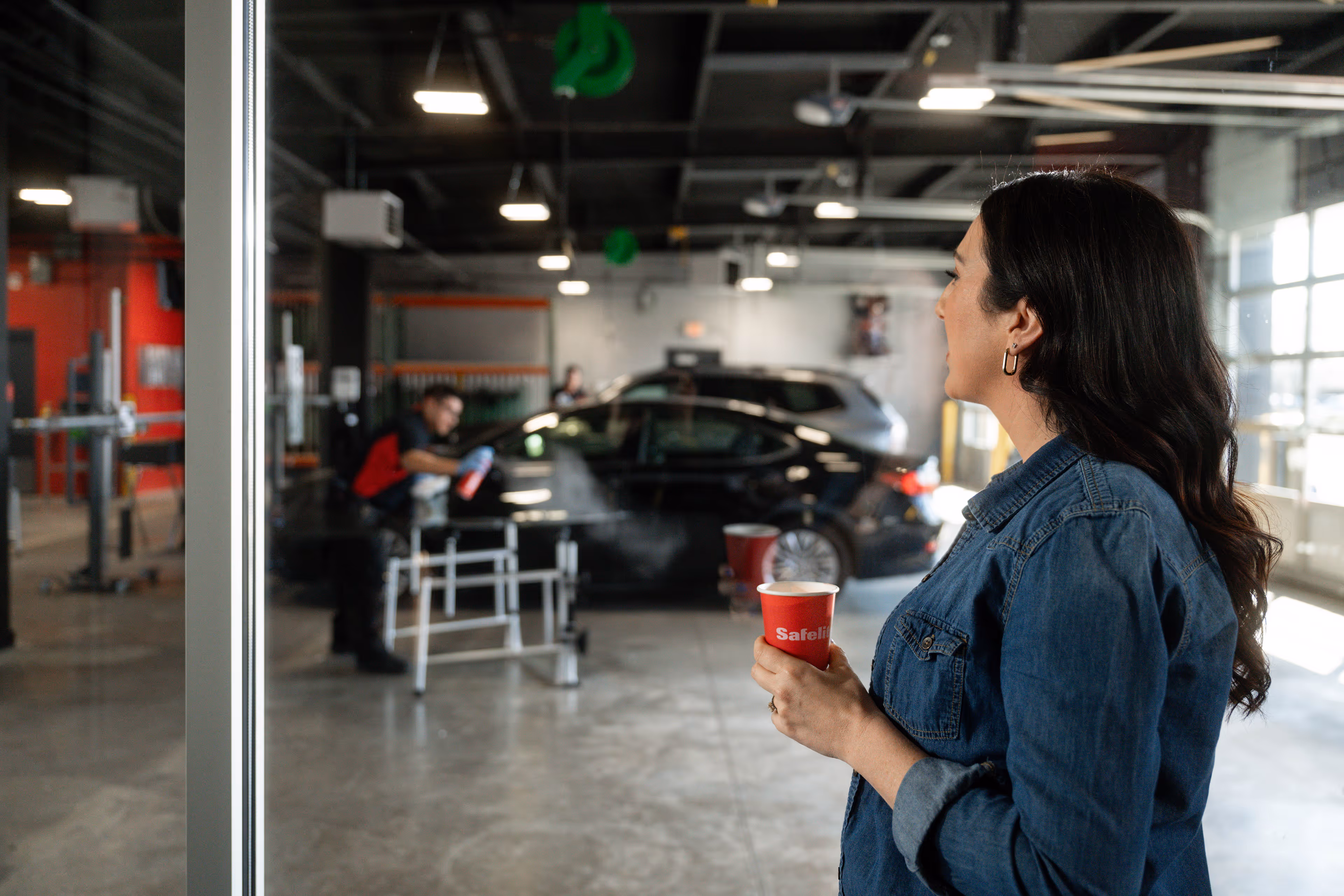 Woman in denim jacket holding a steaming red cup while looking into an auto repair shop where a worker is cleaning a black car.