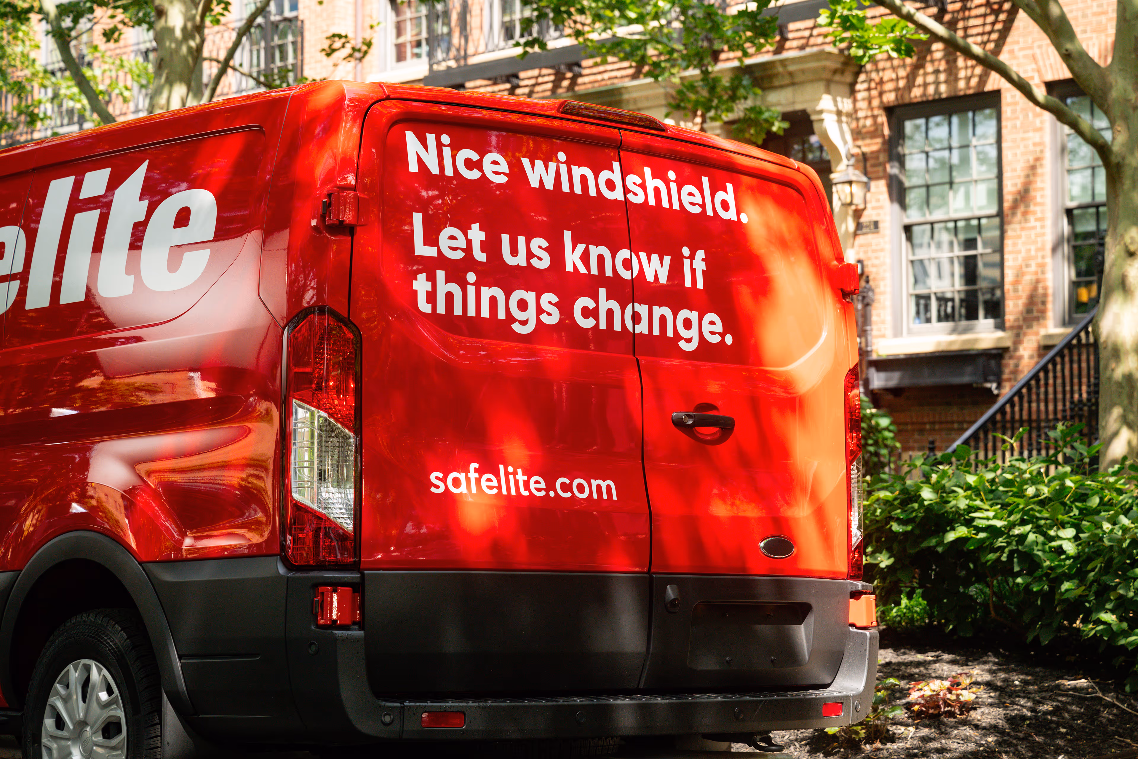 Red Safelite van with white text reading: 'Nice windshield. Let us know if things change.' parked near a brick building with green shrubs and trees.
