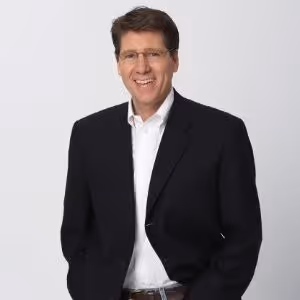 Smiling man wearing glasses, a black blazer, and white shirt against a plain light background.