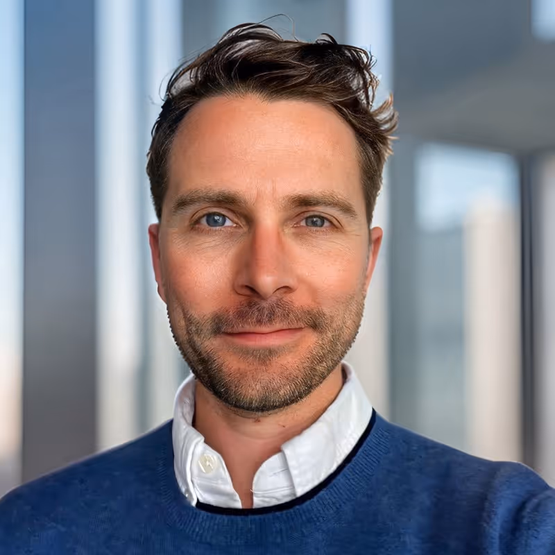 Close-up portrait of a man with blue eyes, brown hair, and light beard wearing a white shirt and blue sweater.
