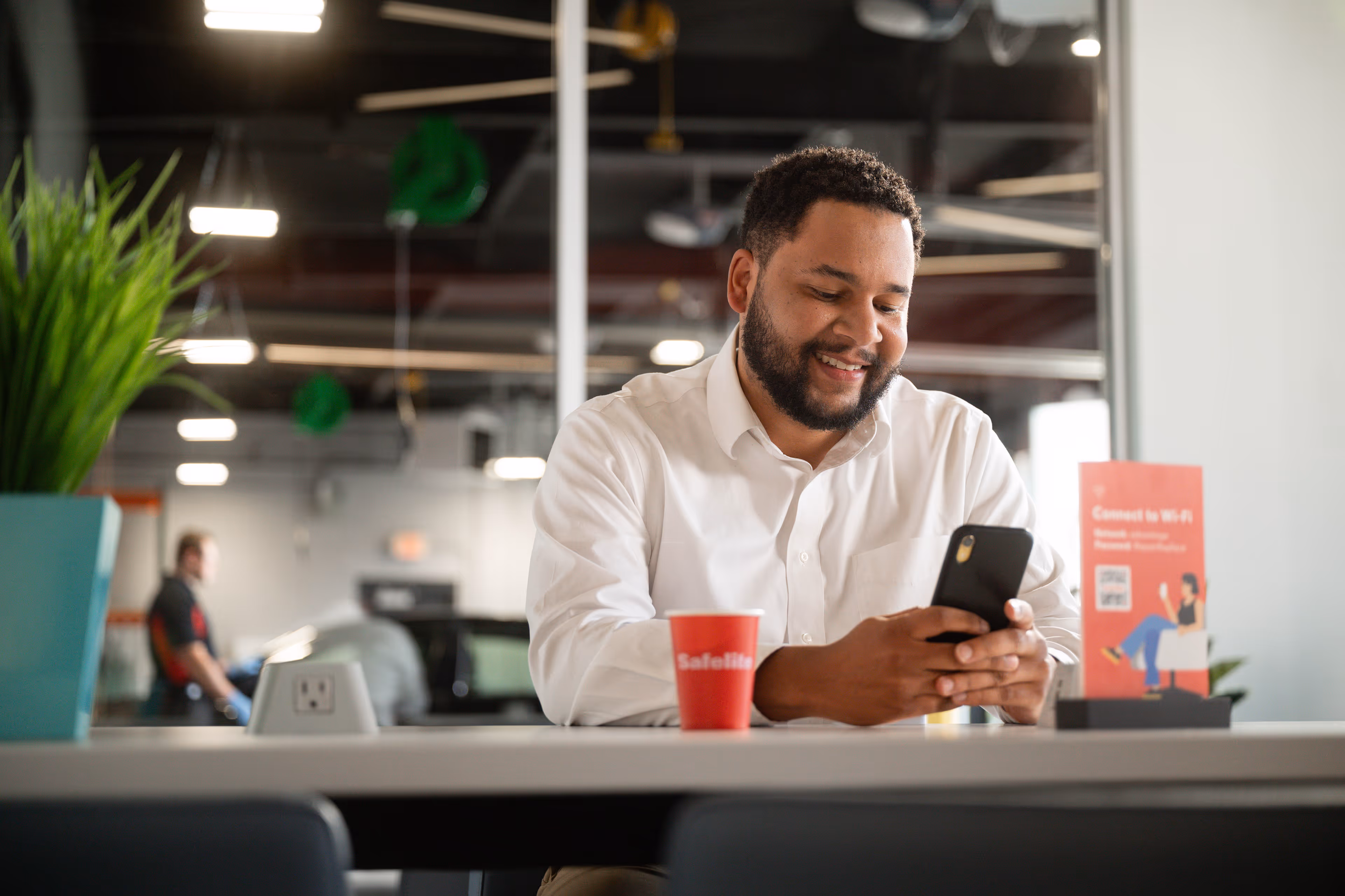 Smiling man in white shirt looking at his smartphone while sitting at a table in a modern office lounge.