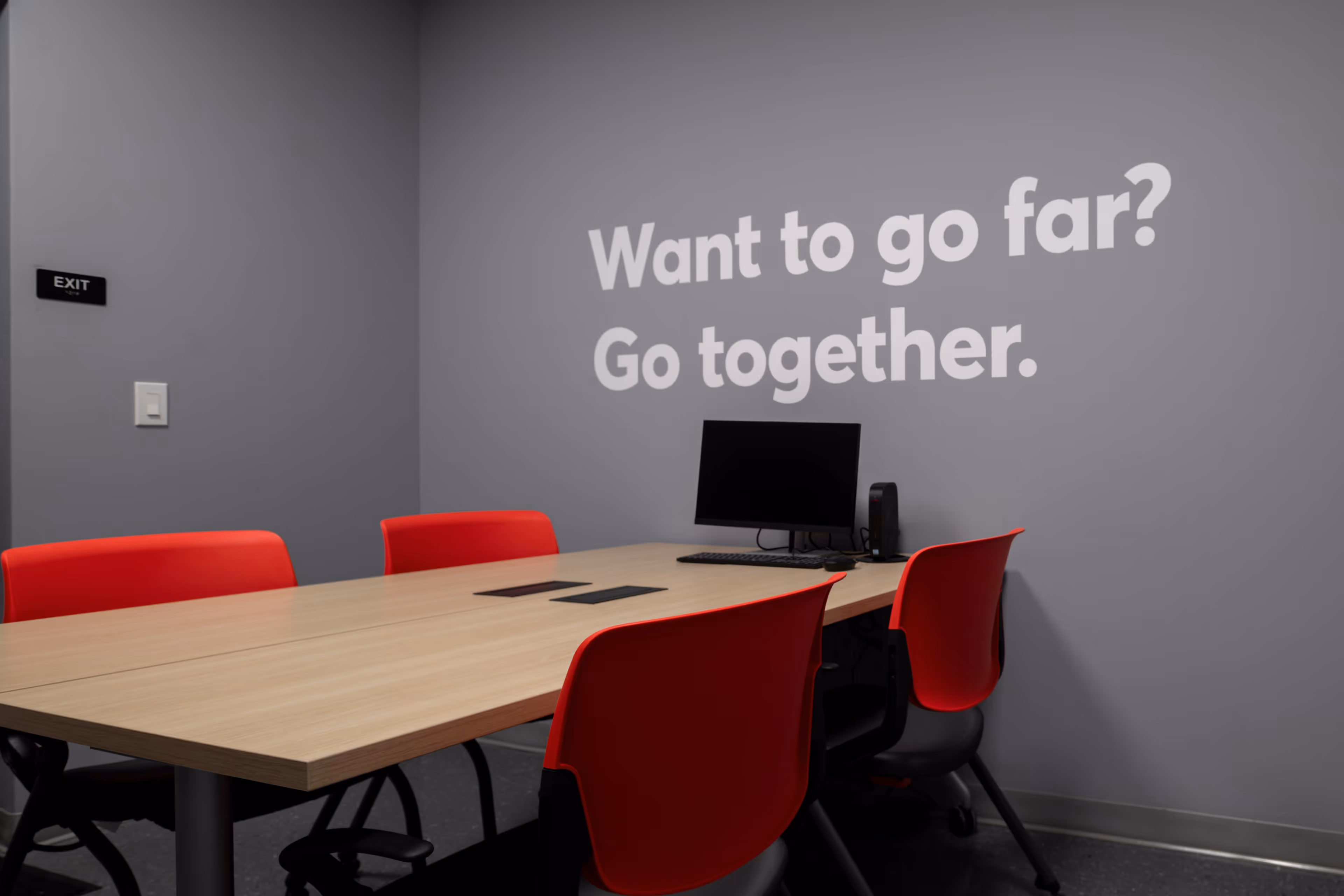 Conference room with a wooden table, four red chairs, a computer, and a wall text that reads 'Want to go far? Go together.'