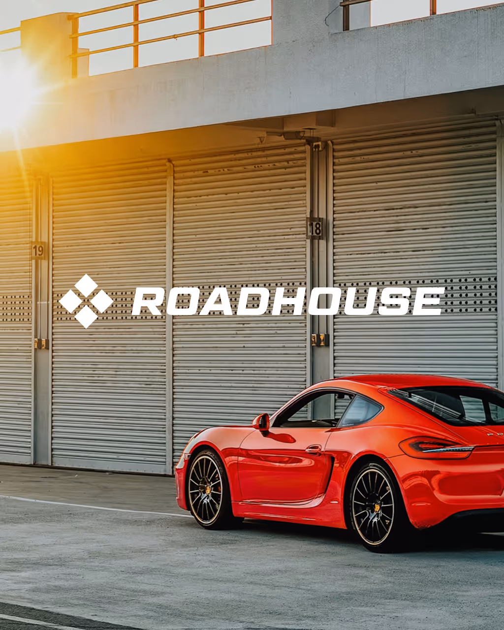 Red sports car parked in front of closed metal garage doors with sunlight shining from the left and the Roadhouse logo overlaid.
