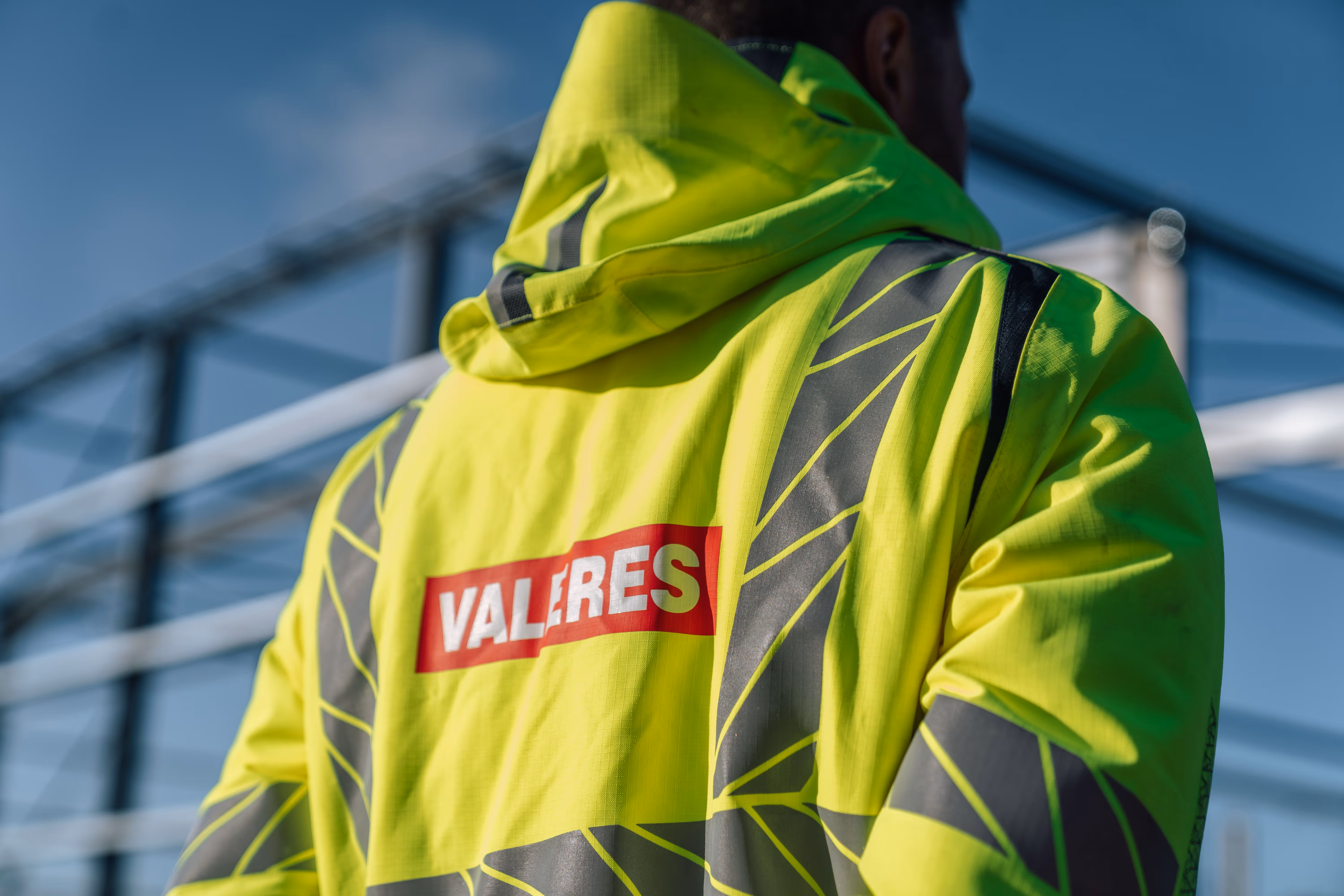 Person wearing a high-visibility yellow safety jacket with reflective black patterns and a hood, branded with a red and white 'VALERES' logo on the back.