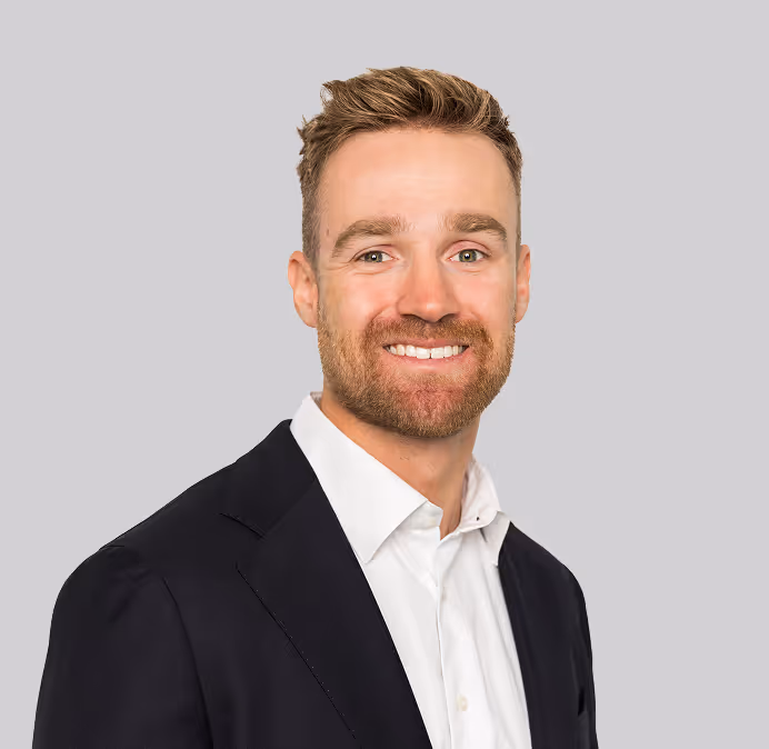Smiling man with light brown hair and beard wearing a white shirt and black blazer against a gray background.