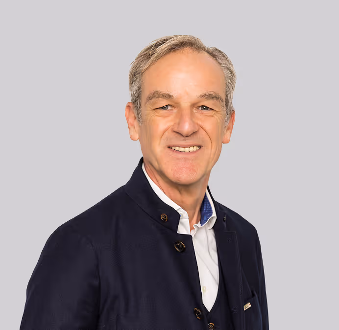 Smiling middle-aged man with gray hair wearing a dark jacket and white shirt against a light gray background.