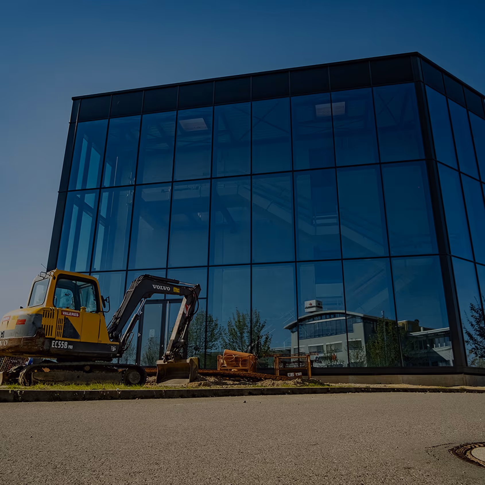 Yellow Volvo EC55B excavator parked beside a modern glass building reflecting nearby structures on a clear day.