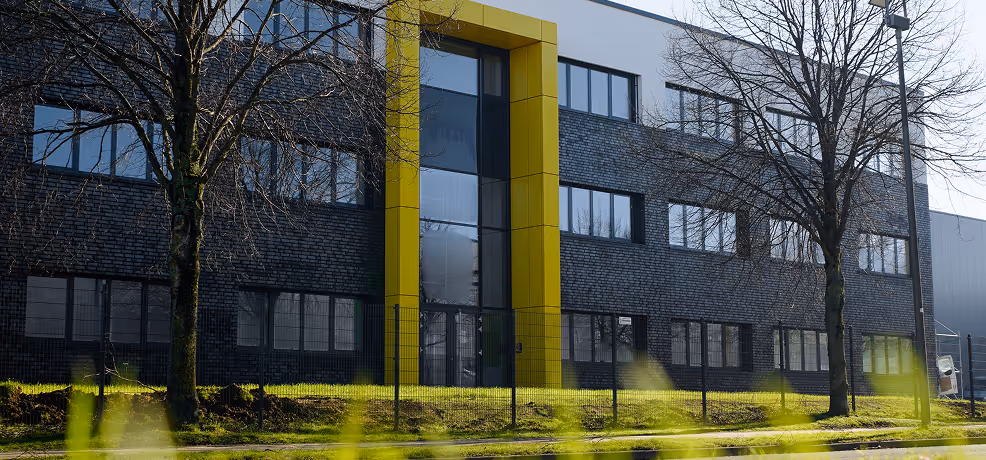 Modern dark brick building with large windows and a tall yellow-framed glass entrance, flanked by leafless trees and a metal fence.