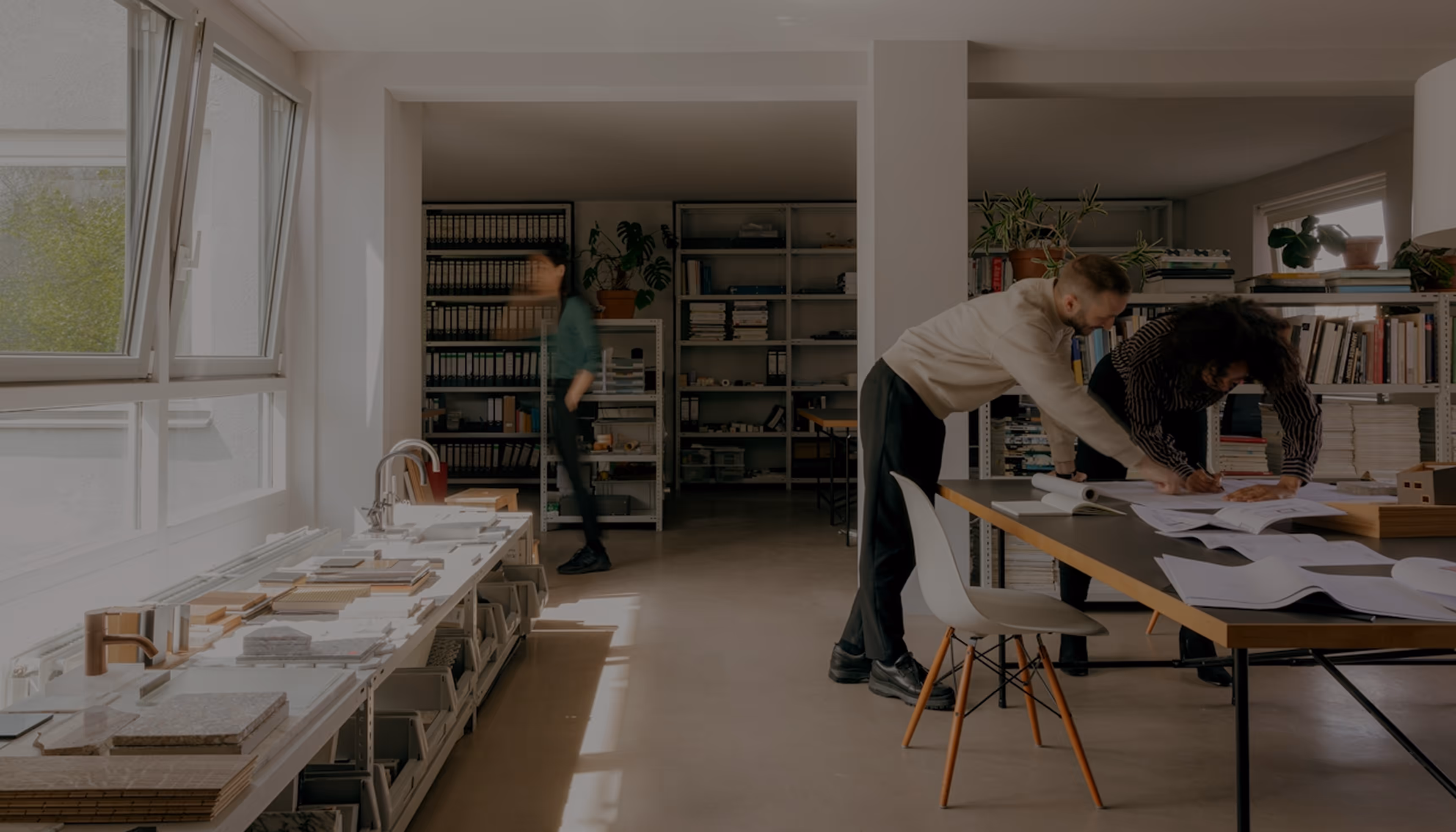 Two people leaning over a table reviewing architectural plans in a bright, modern office with shelves and samples.