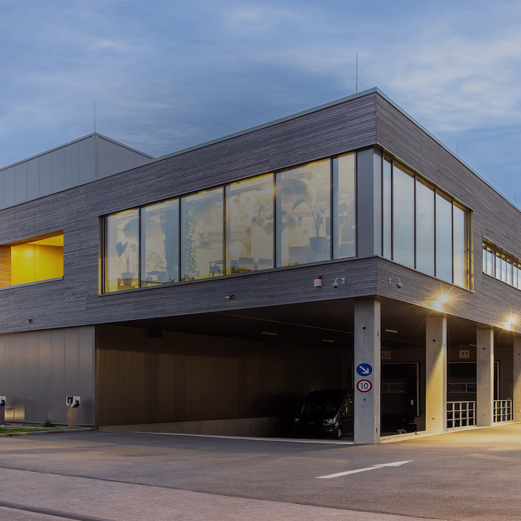 Modern office building with large windows illuminated inside and parking area underneath at dusk.