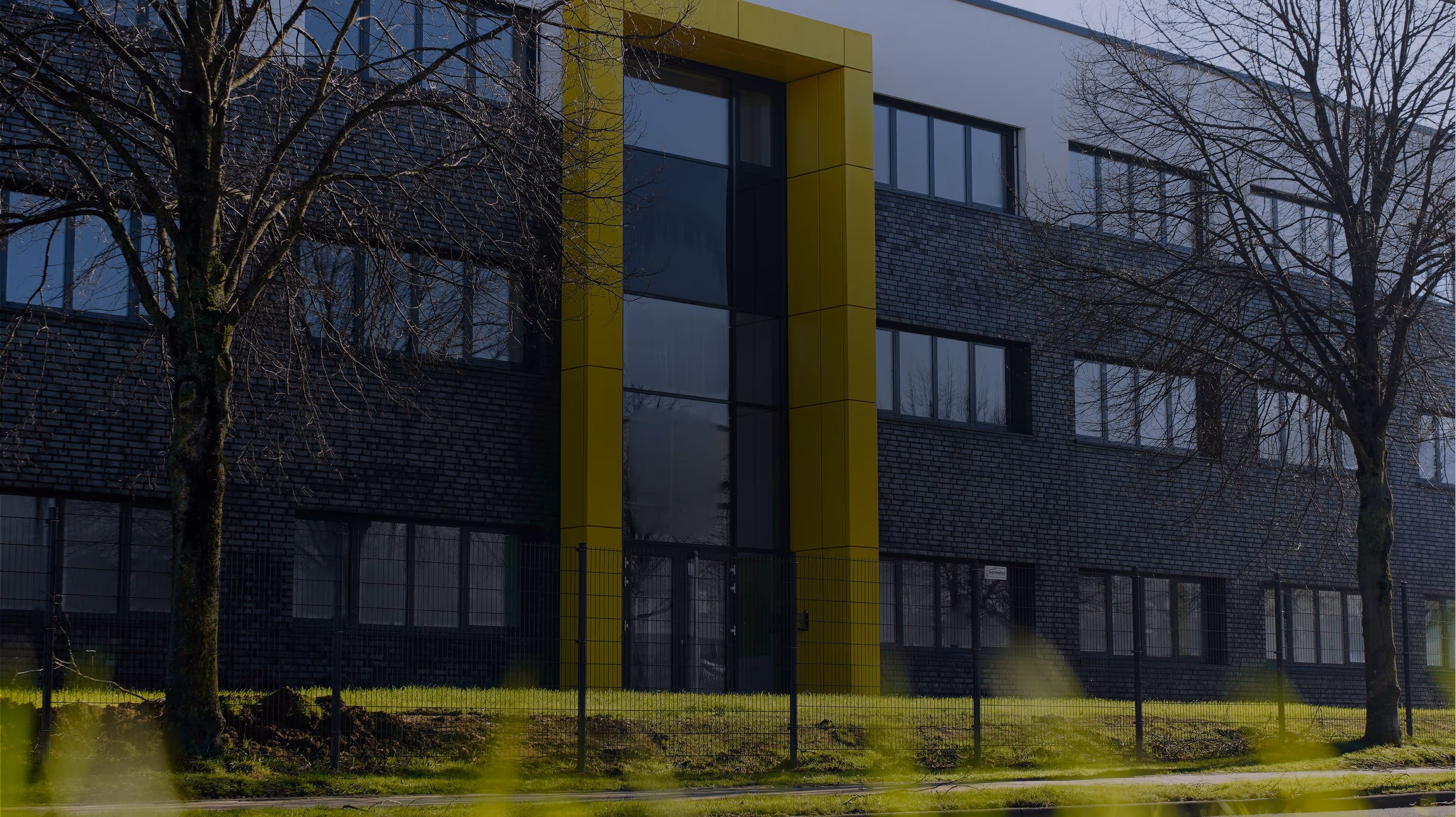Modern office building with dark brick facade and tall yellow-framed glass entrance surrounded by leafless trees.