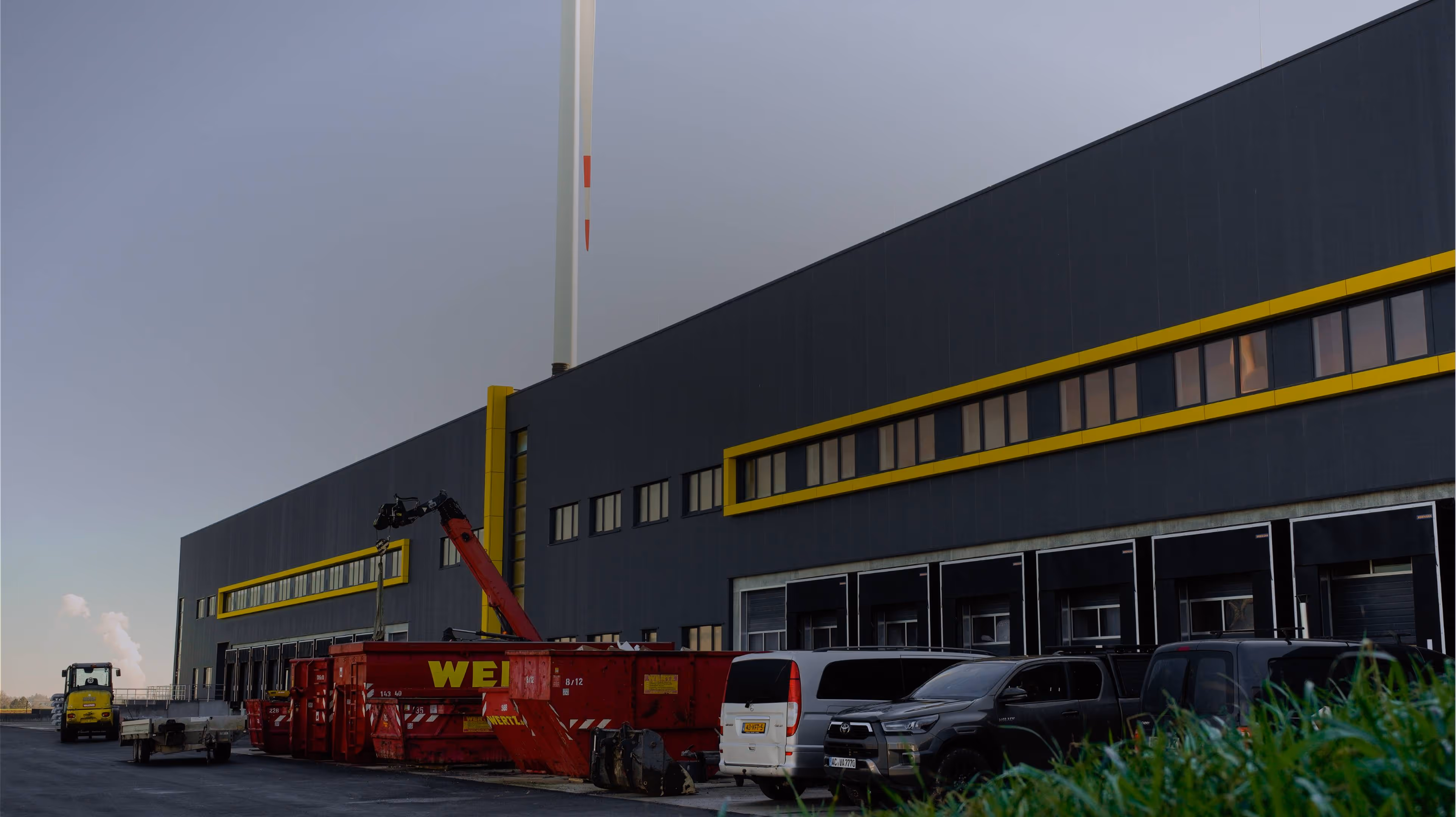 Exterior of a large industrial building with yellow-trimmed windows, parked vehicles, and a red container with crane equipment.