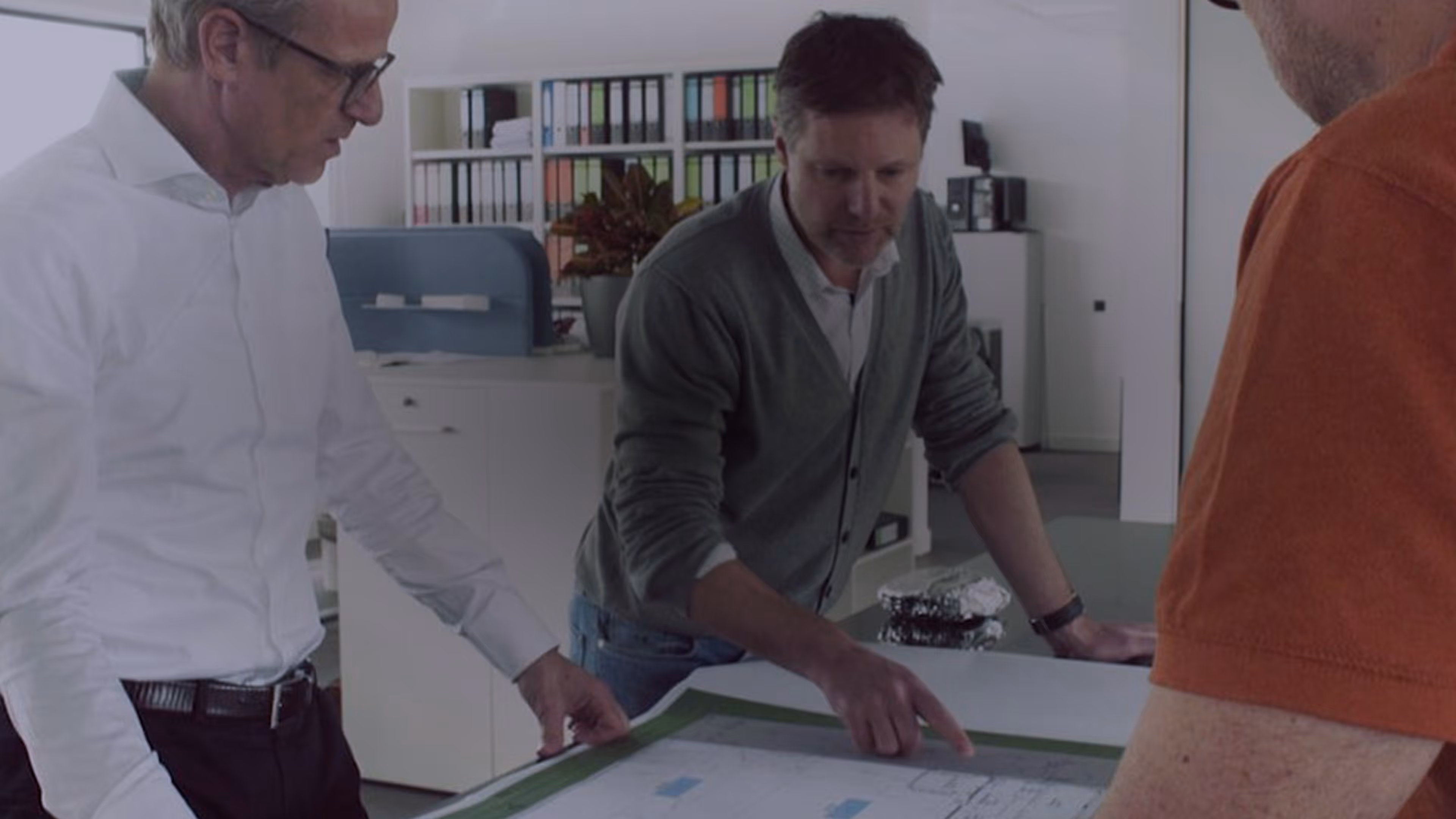 Three men in an office reviewing and discussing architectural plans spread on a table.