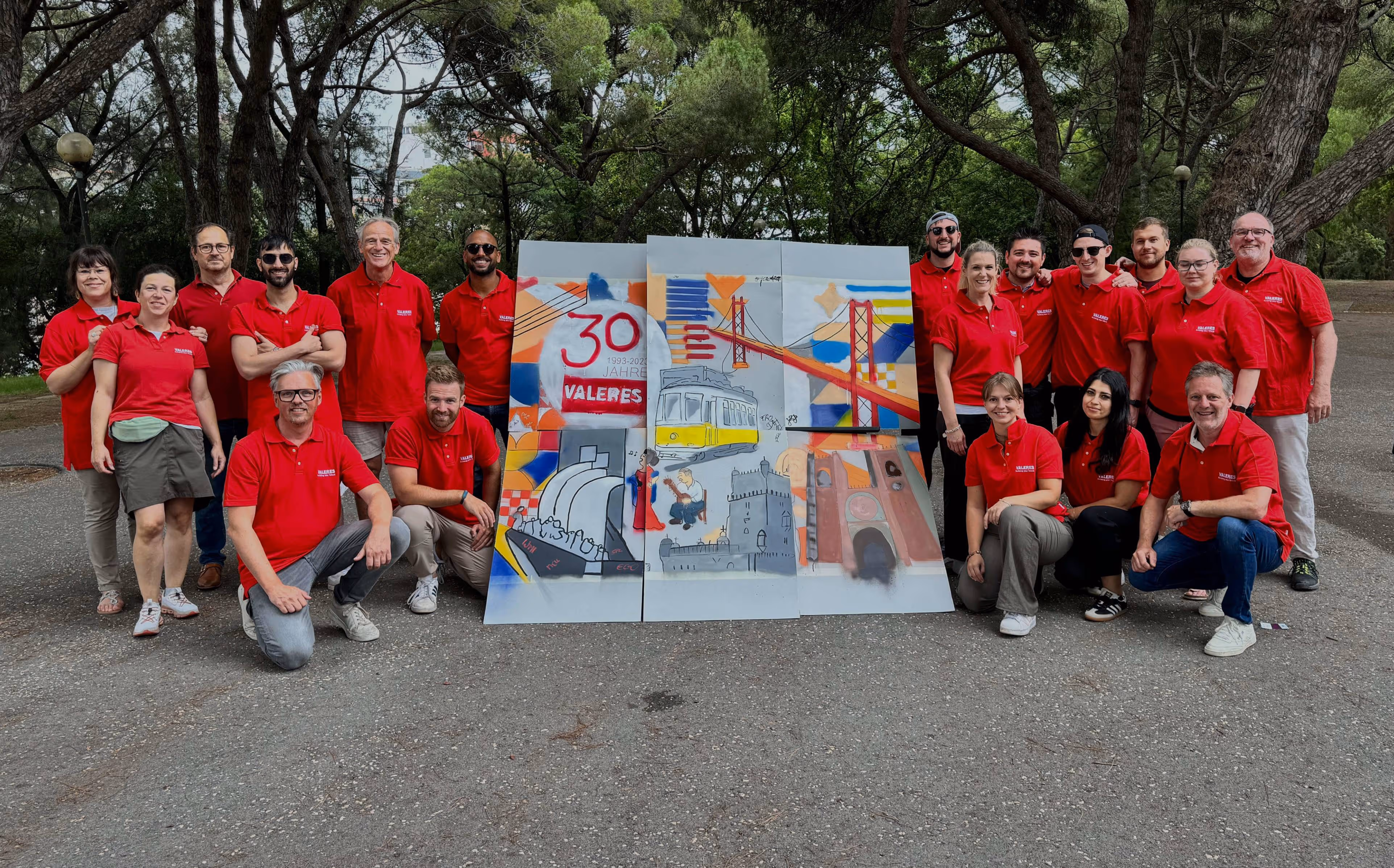 Group of people wearing matching red polo shirts posing outdoors around a three-panel painted artwork depicting a yellow tram, a red bridge, and other colorful city elements.