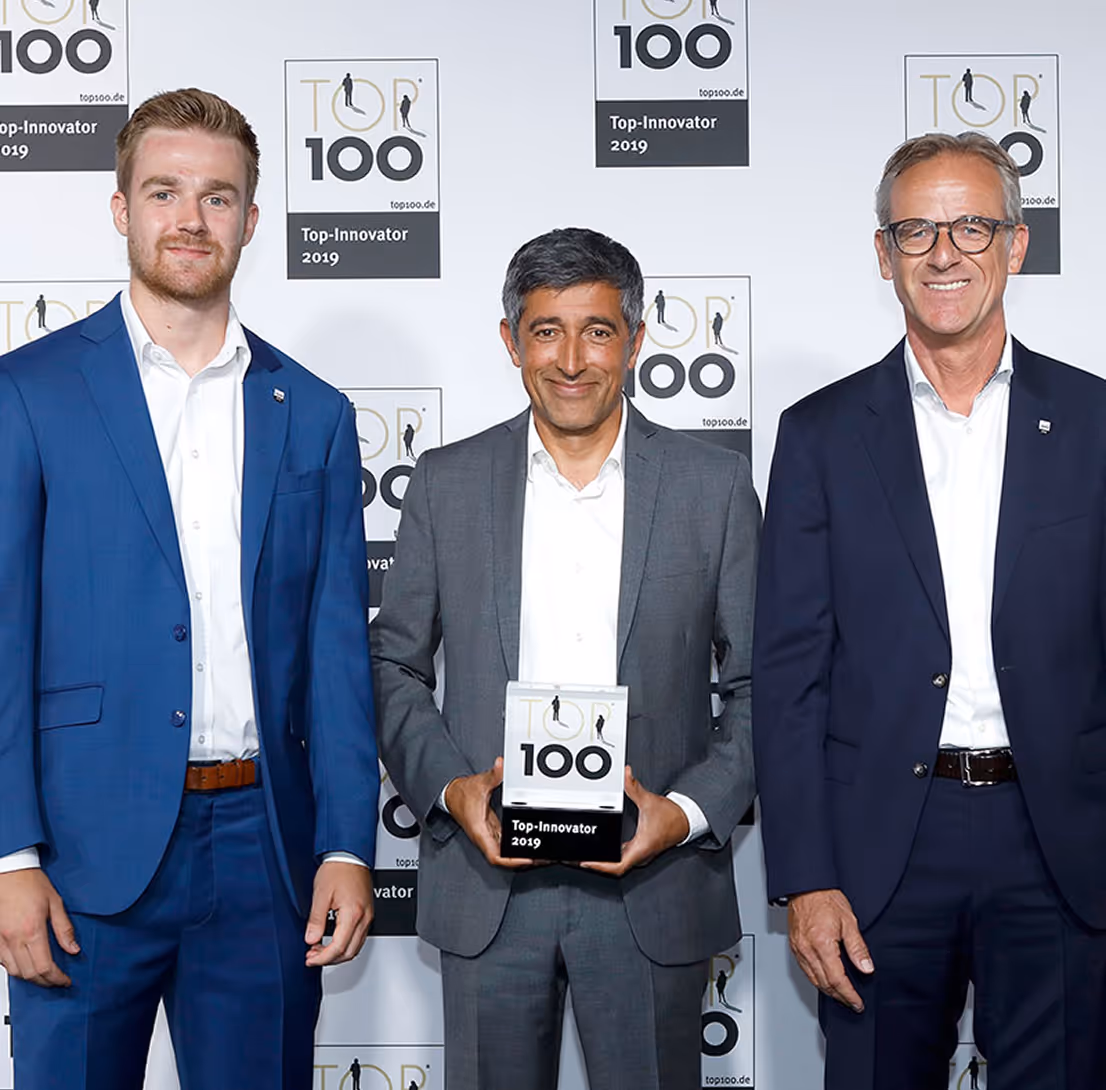 Three men in suits standing in front of a Top 100 Innovator 2019 backdrop, the man in the center holding a Top 100 Innovator award.